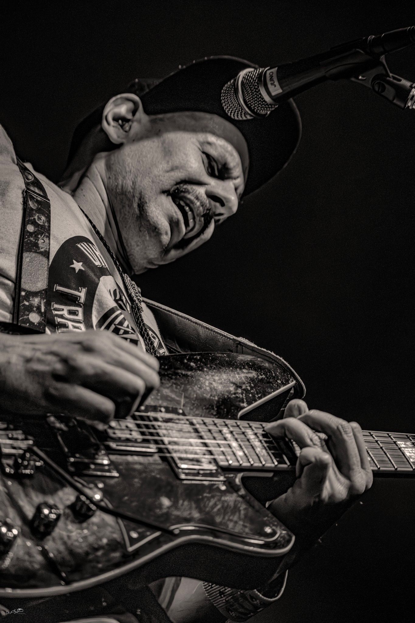 Guitarist playing a guitar, lit by stage lights, wearing a hat, focused expression.