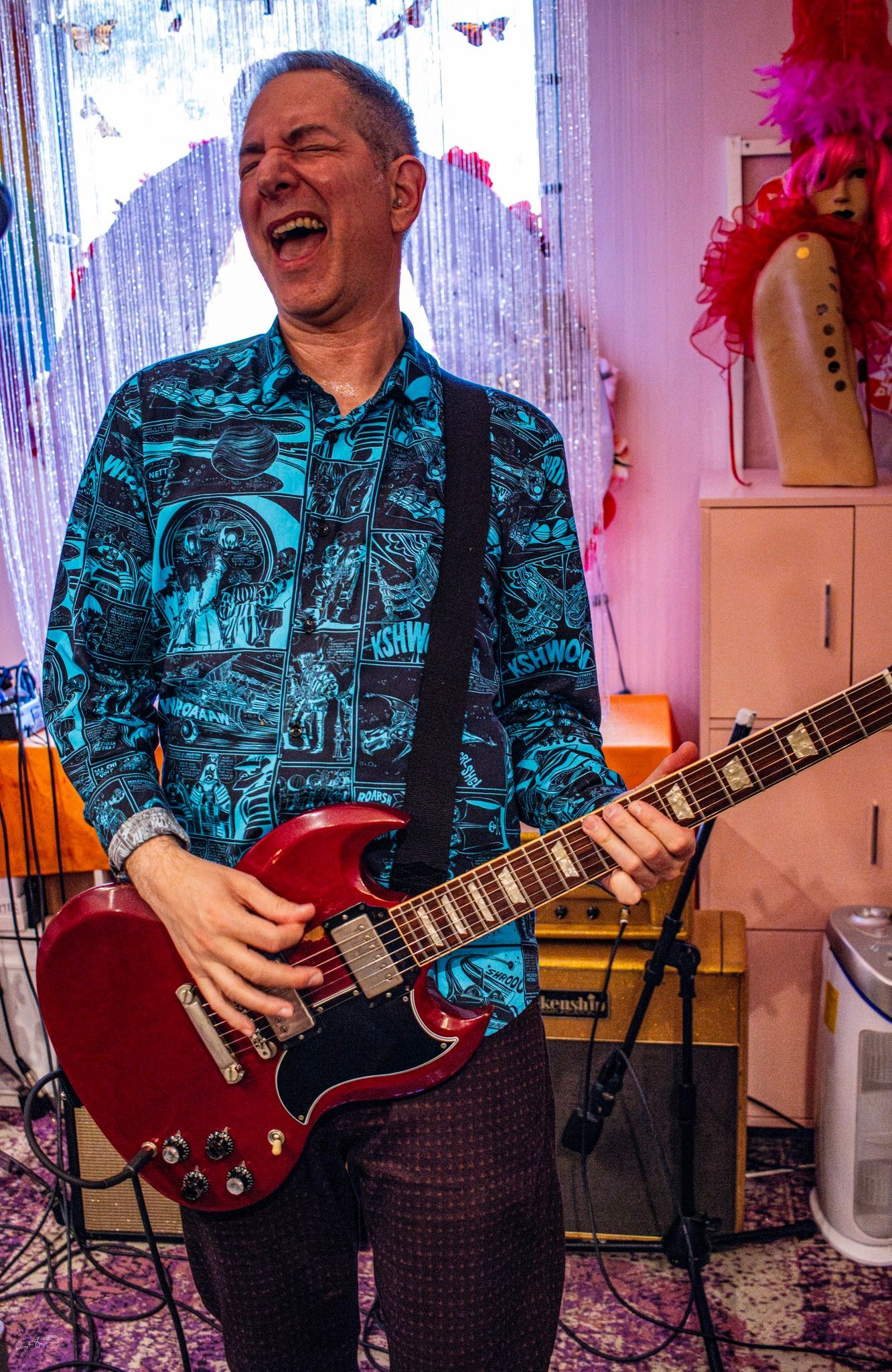 Man playing red electric guitar, smiling, wearing blue patterned shirt, indoors.