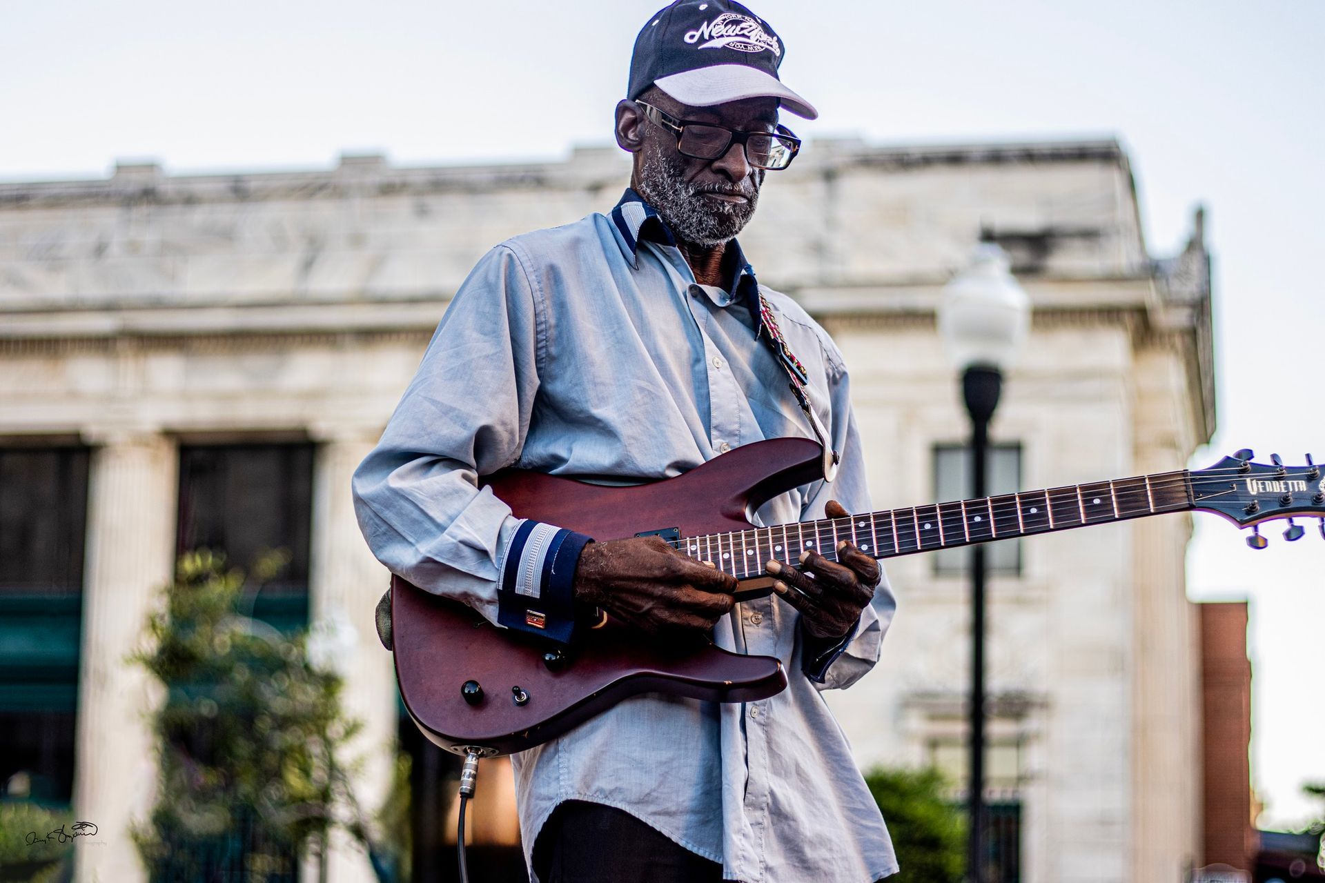 Man playing electric guitar outdoors. Wearing a light blue shirt, cap, and glasses. A building is in the background.