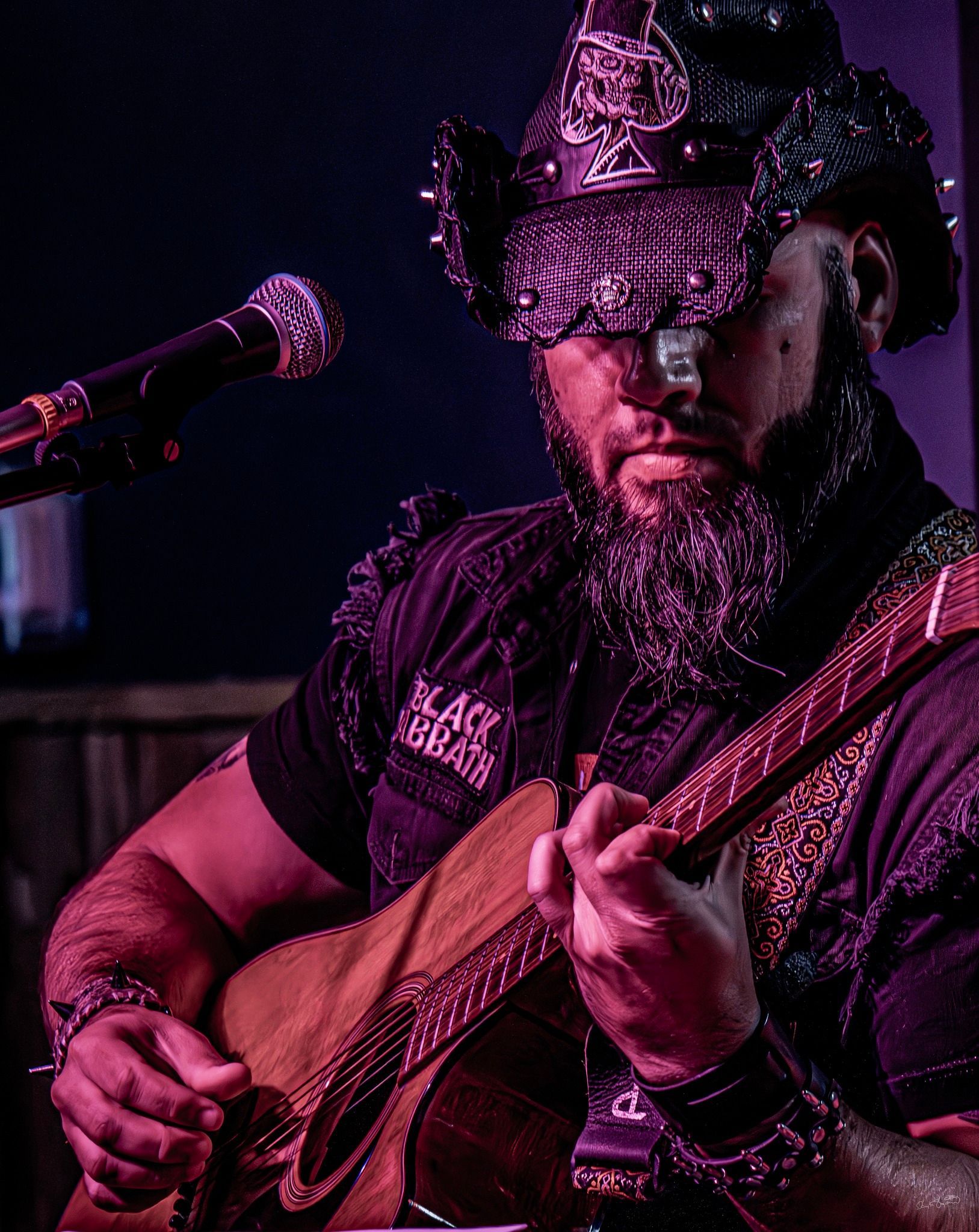 Man in embellished hat and clothing playing a stringed instrument, lit in purple, in a dimly lit setting.