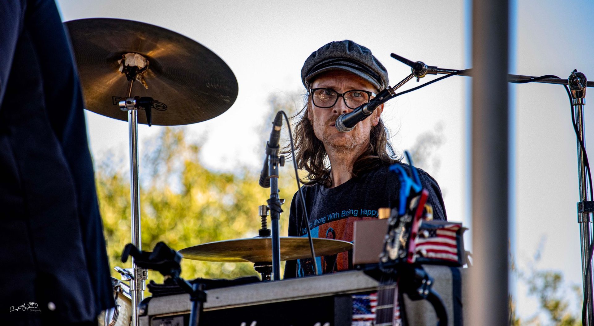 Drummer wearing a cap and glasses plays outdoors, with a dark shirt, in front of a blurred background.