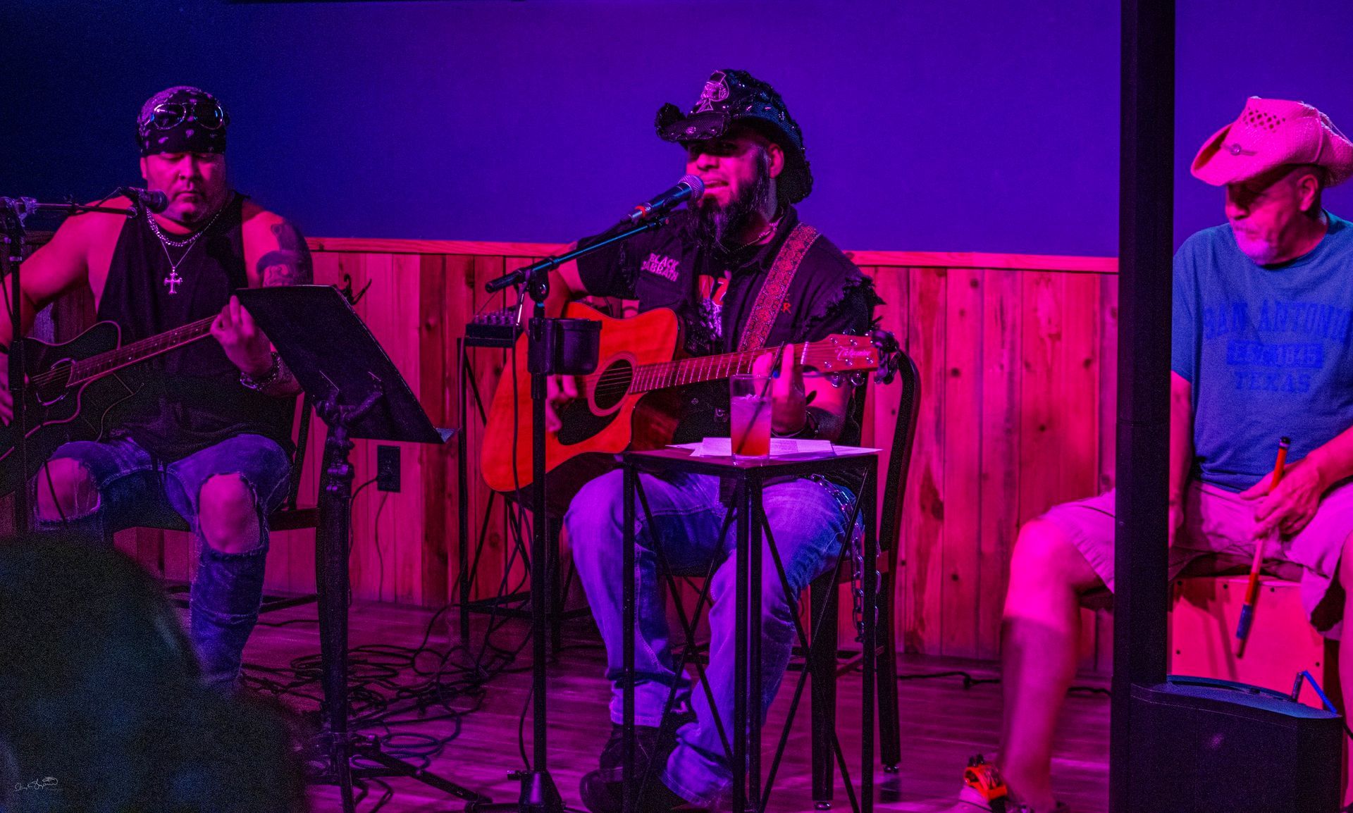 Three musicians performing on a stage lit with purple and red light. One plays guitar, another, seated, sings into a microphone, and the third sits to the side.