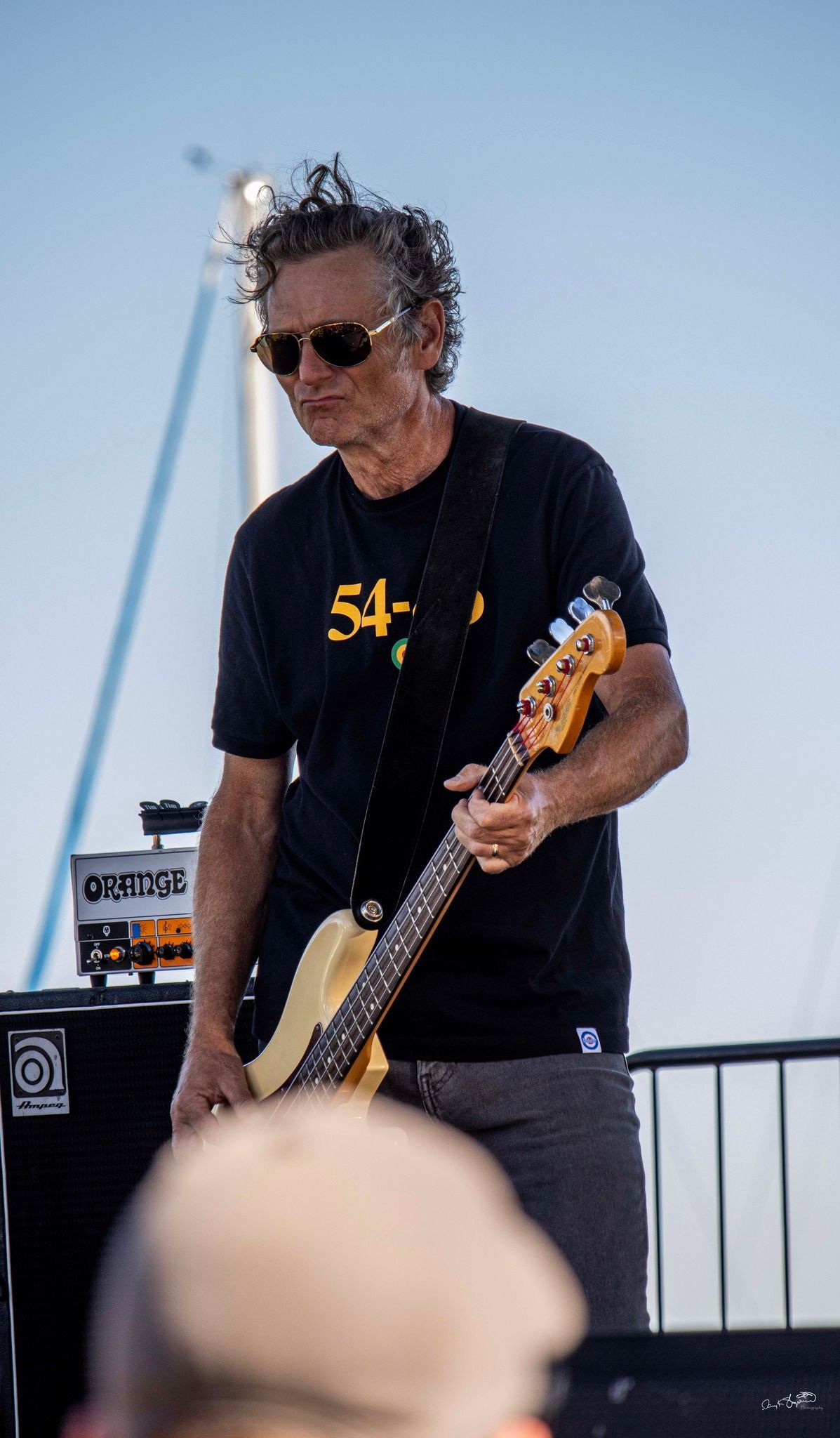 Man playing bass guitar on outdoor stage, wearing sunglasses and black t-shirt with “54-40” logo.