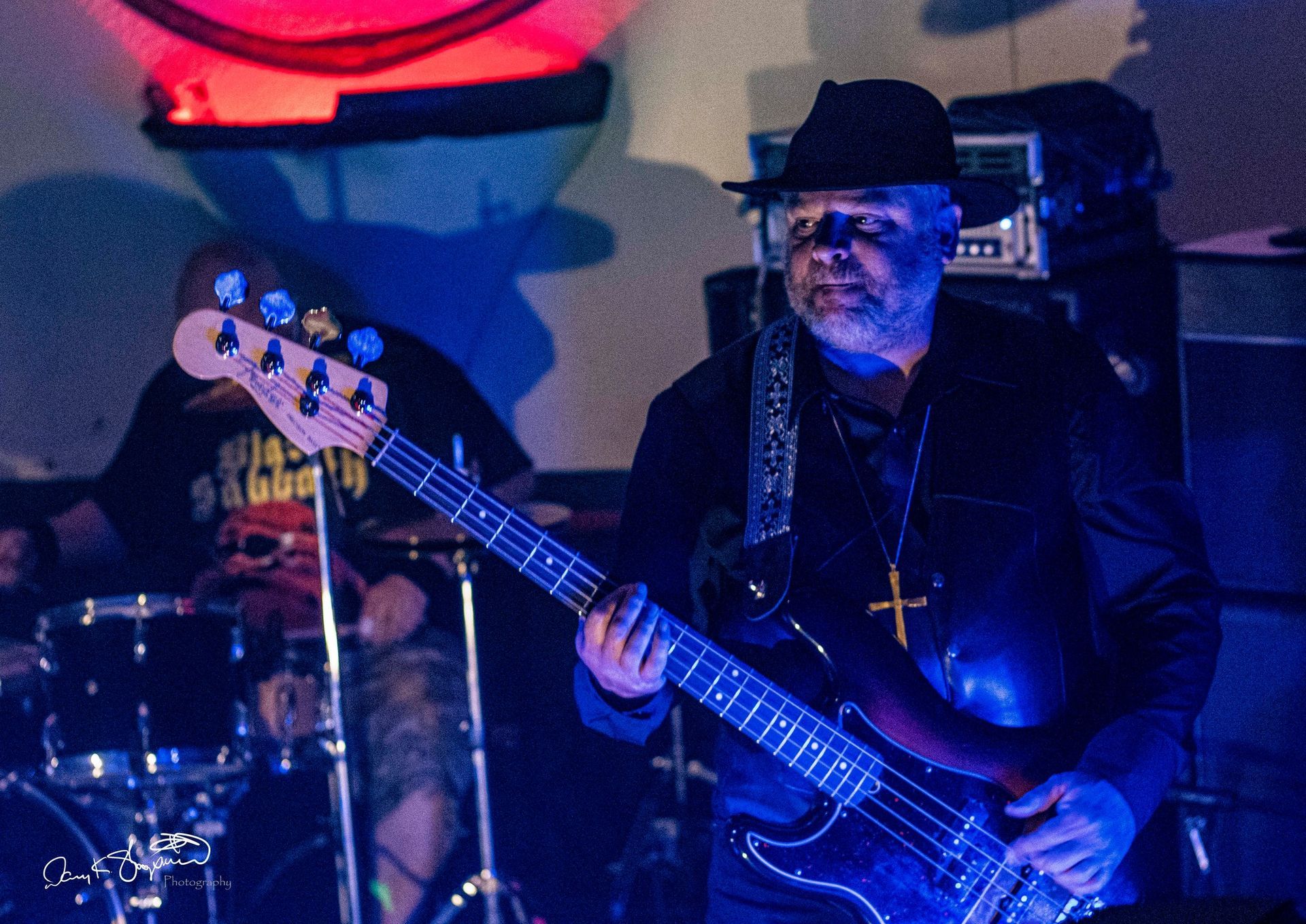 A bassist in a fedora plays electric bass on stage, illuminated by blue and red light. A drummer is in the background.