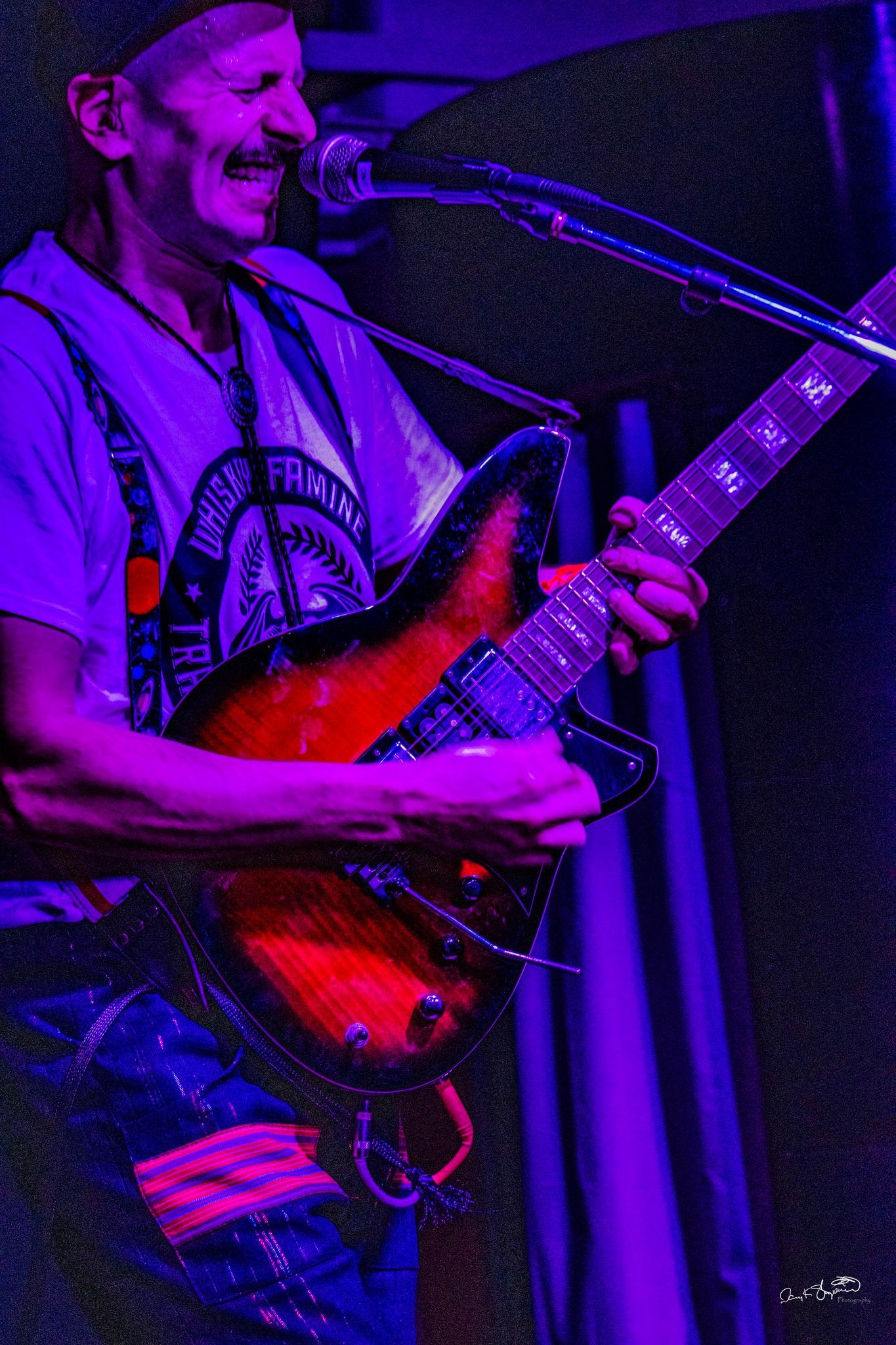 Man playing guitar onstage, smiling, under blue stage lighting. Sunburst electric guitar, dark background.