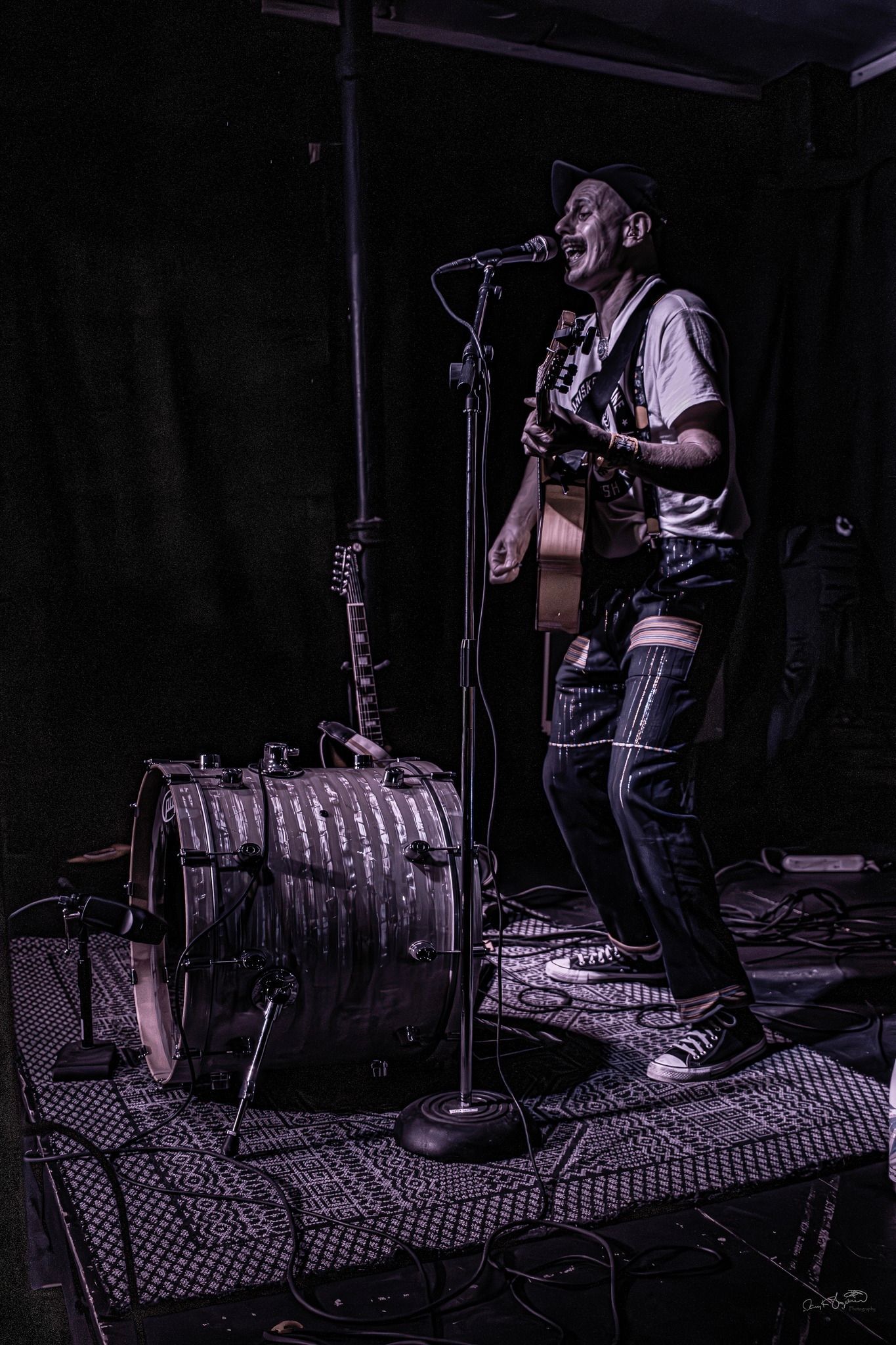A musician performs onstage, playing guitar. Dark setting, wearing a hat, mic stand nearby.