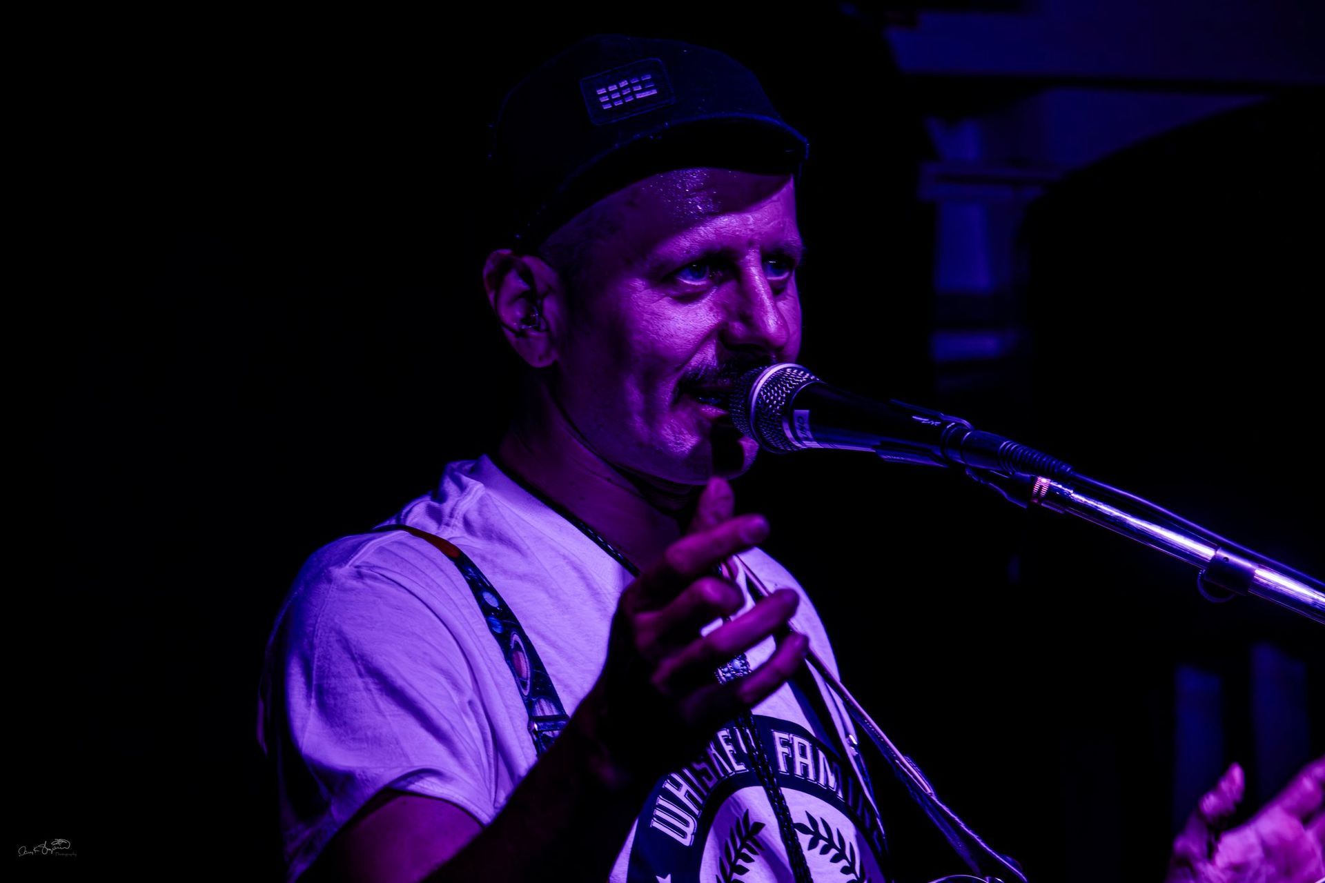 Man singing into a microphone, lit with purple light. He wears a hat and suspenders, gesturing with his hands.