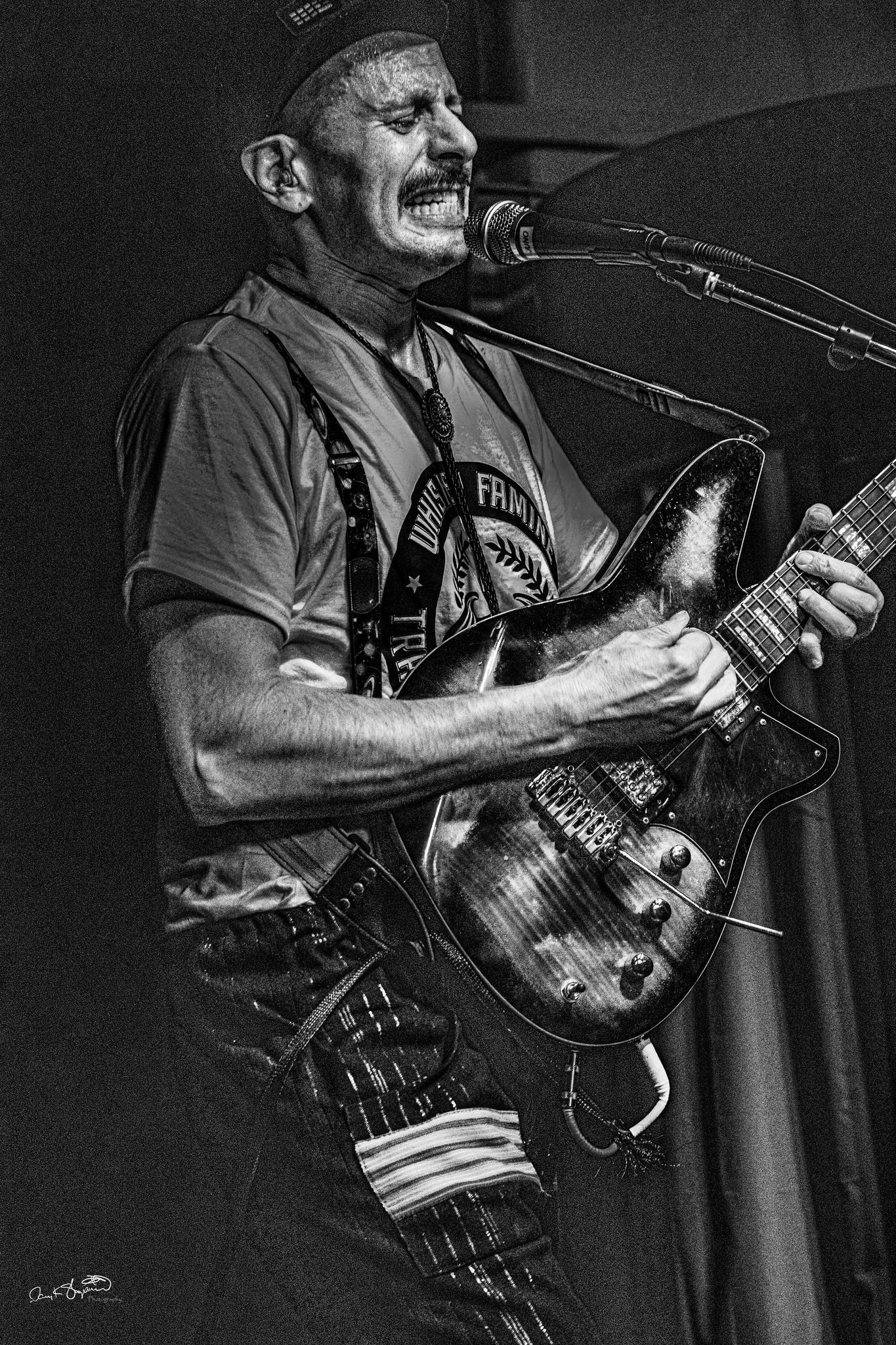 Musician playing guitar, stage performance. Dark, with suspenders and microphone.