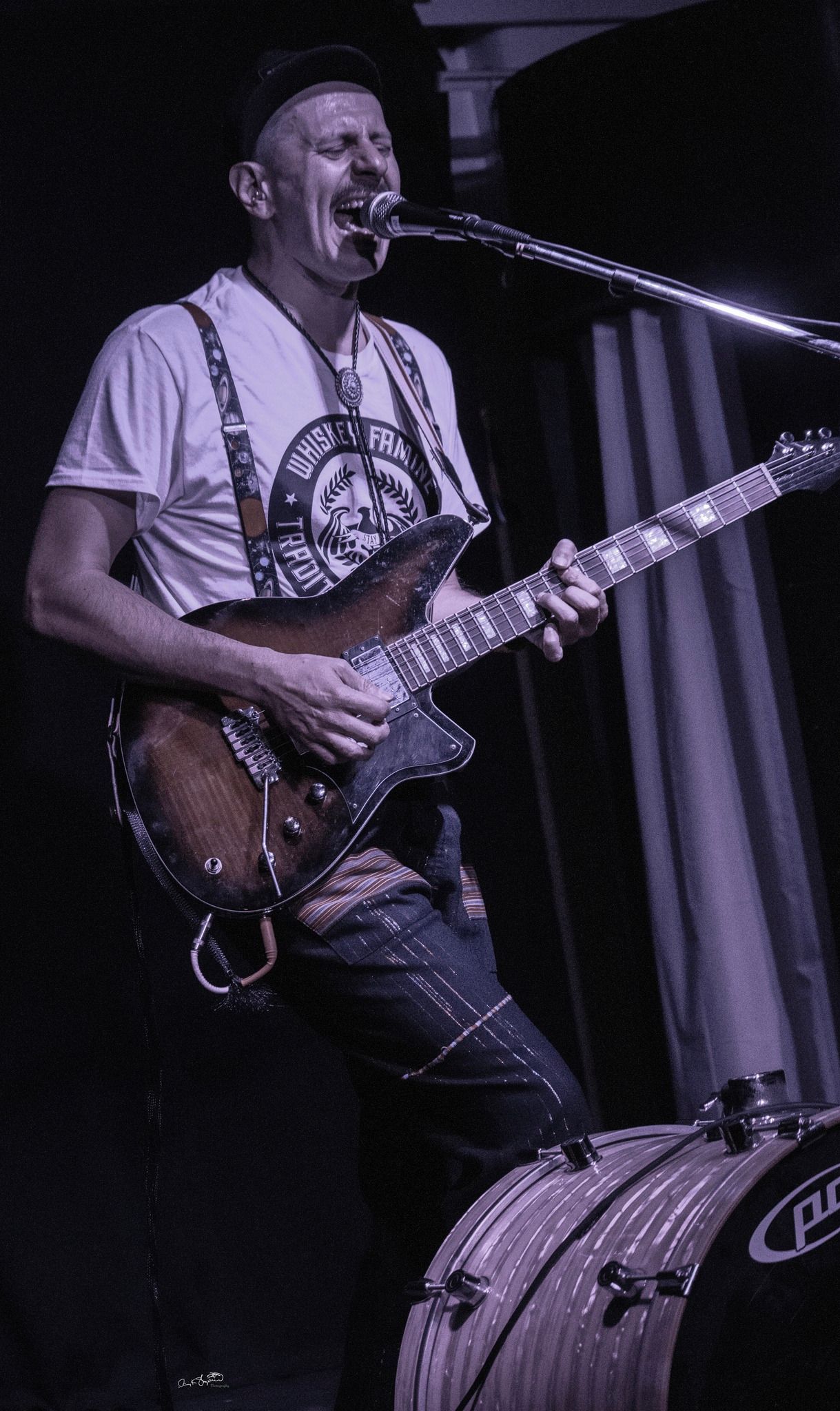 Man playing guitar and singing at a concert, wearing a hat, suspenders, and paint-splattered jeans.