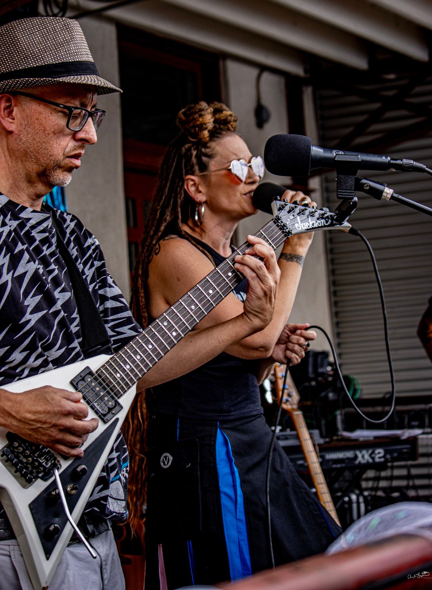 Band performing outdoors: guitarist with white V-shaped guitar, singer with sunglasses and mic.
