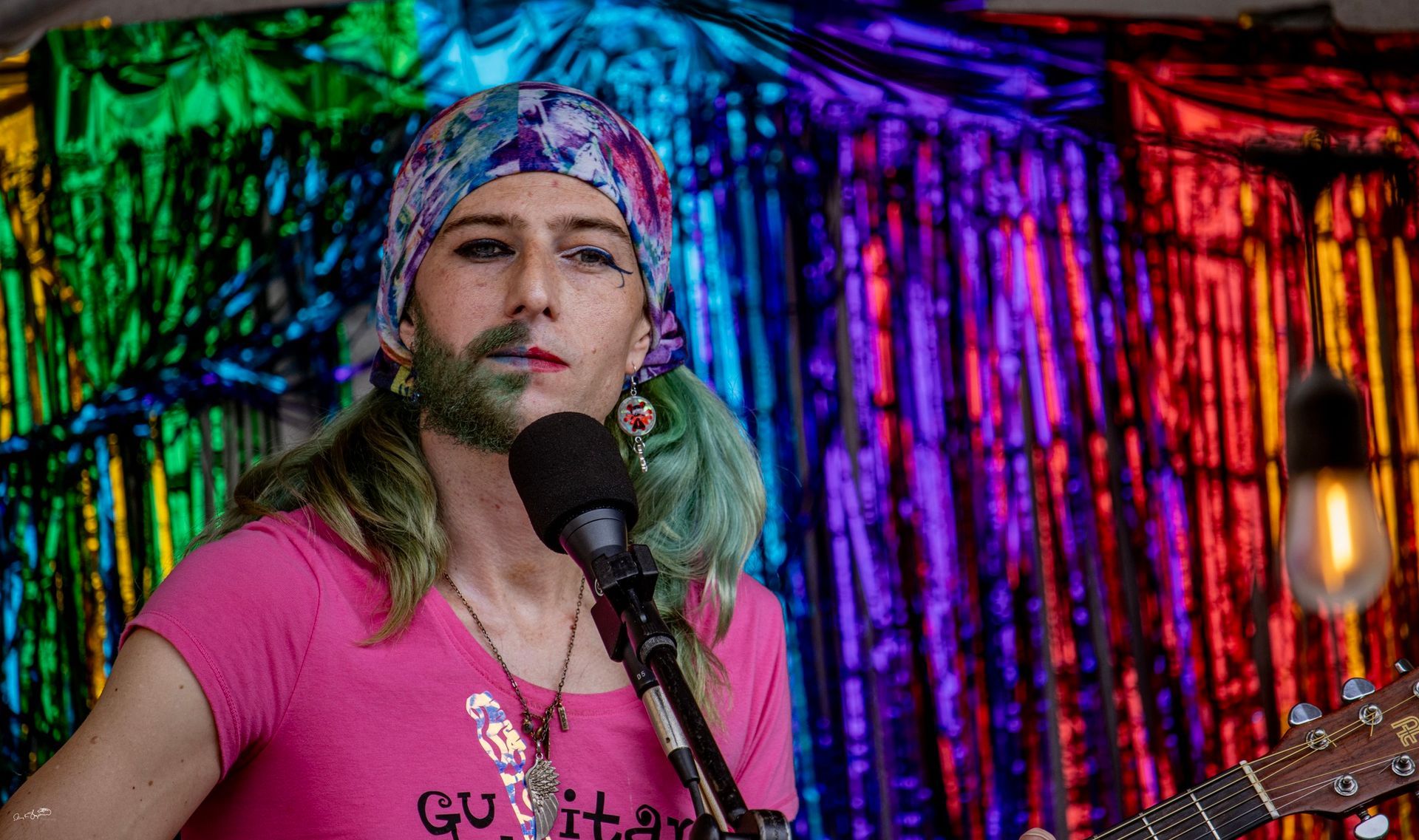 Person with green-tinted beard, pink shirt, playing guitar, singing into microphone. Rainbow backdrop.