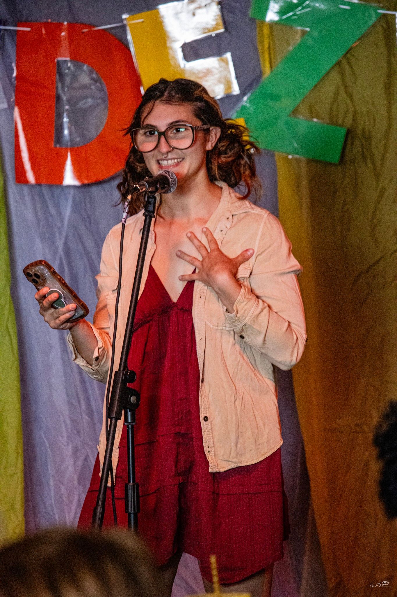 Woman with glasses and a microphone on stage, speaking with hand gestures, holding a phone. Stage background with colored letters.