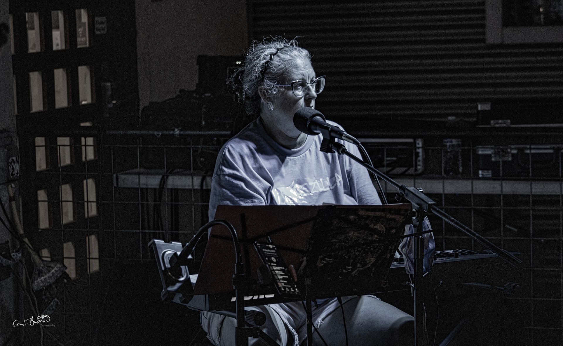 A person with glasses singing into a microphone, sitting at a keyboard in a dimly lit setting.