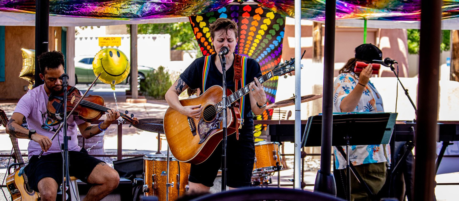 A band performing on an outdoor stage beneath a colorful canopy. Musicians playing guitar, violin, keyboard, and percussion.