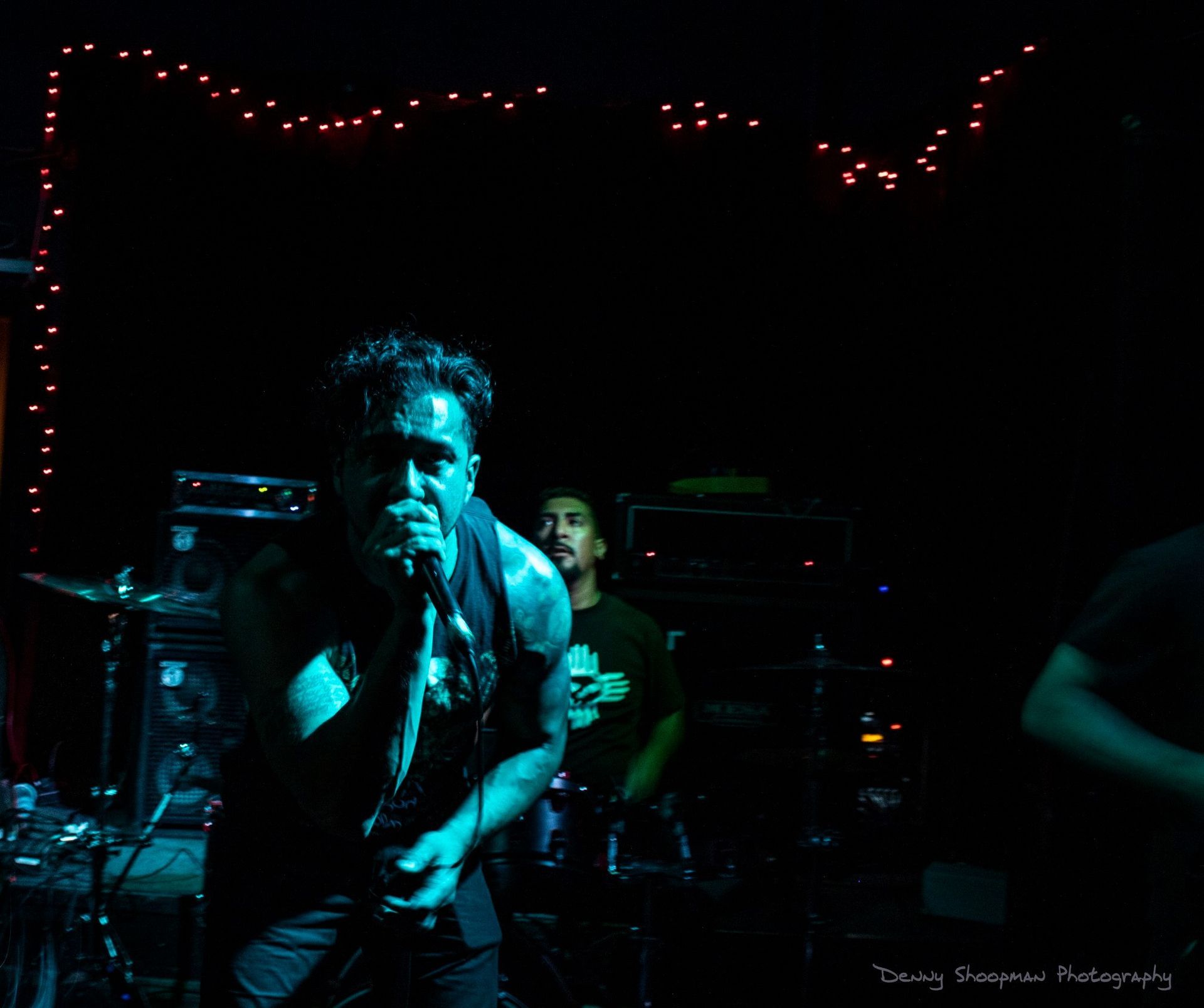 Band performing onstage; vocalist singing into a microphone, lit by blue lights; drummer in the background.