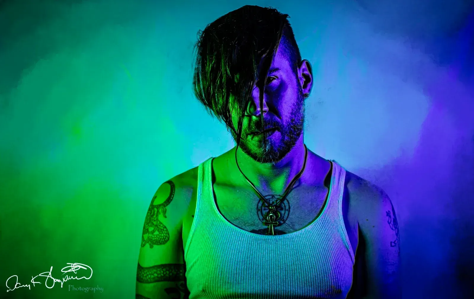 Man with beard and dark, wet hair, lit with green and blue lights. Wearing a white tank top and pendant.