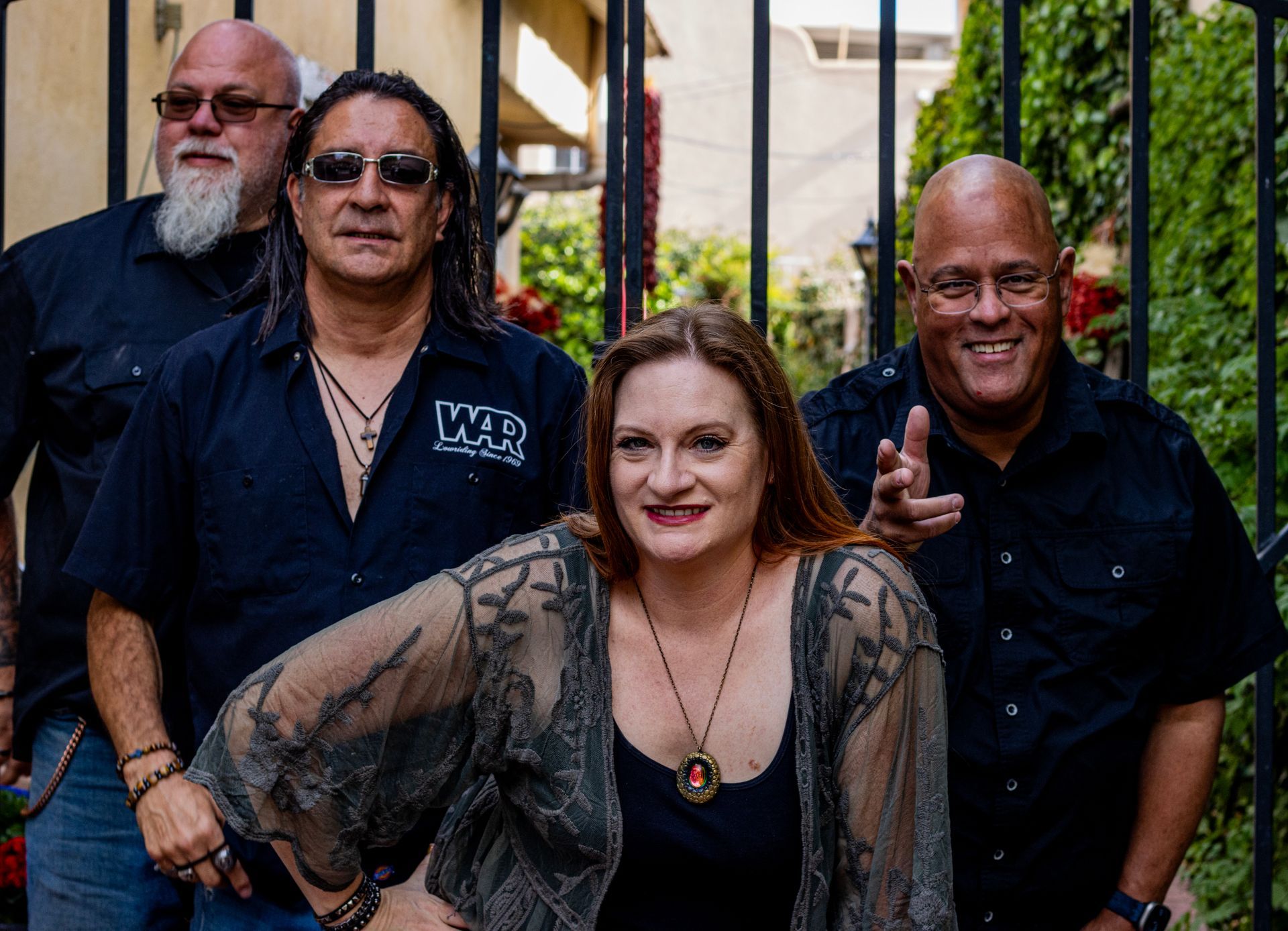 Four band members posing outdoors, woman in front, men behind, smiling. Black clothing, wrought iron gate.