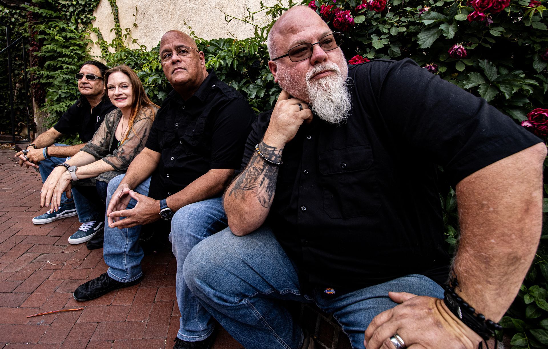 Four people sitting outside on a brick bench. One man has a beard and arm tattoo.