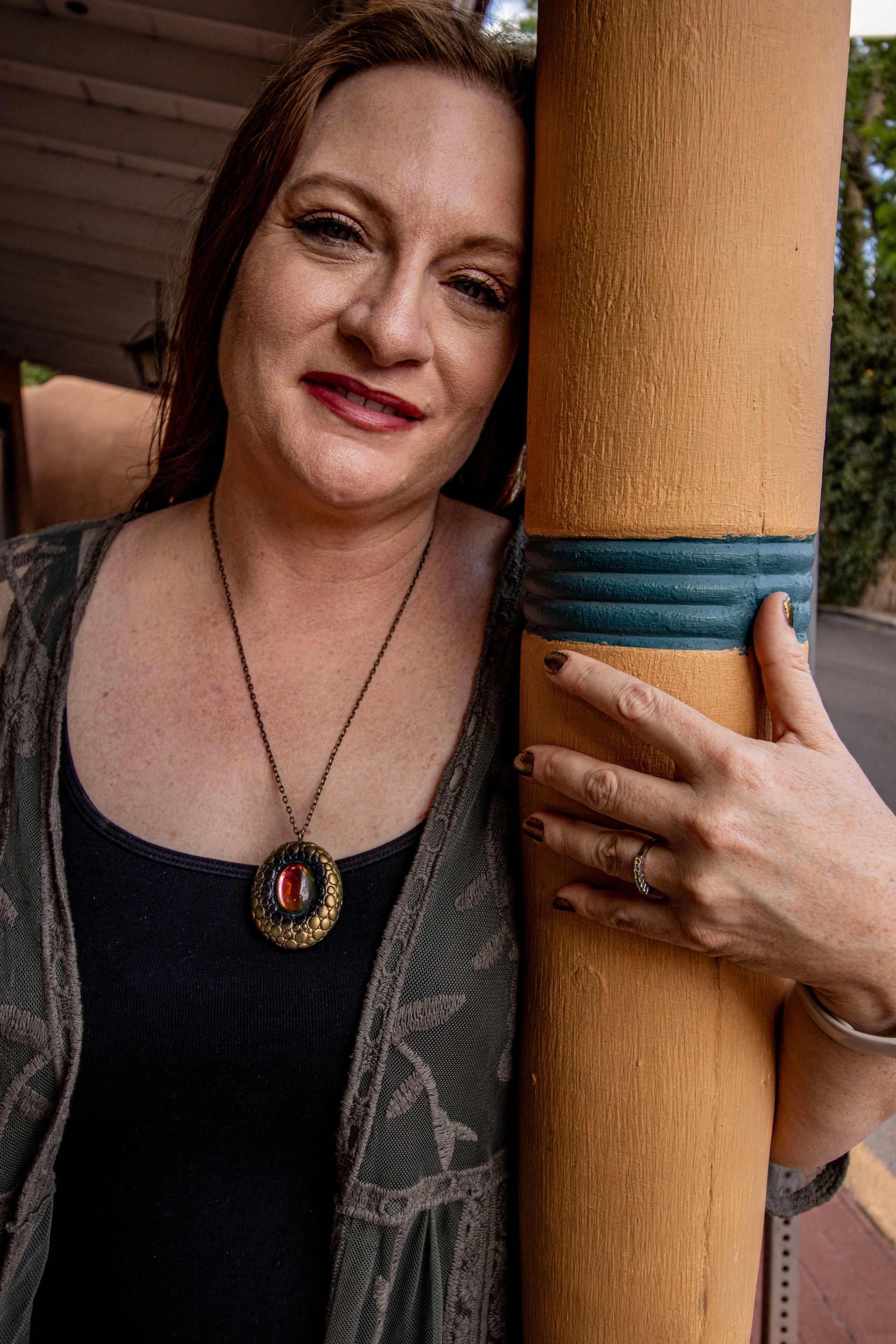 Woman leaning against a tan pillar, wearing a patterned vest and pendant necklace, smiling.