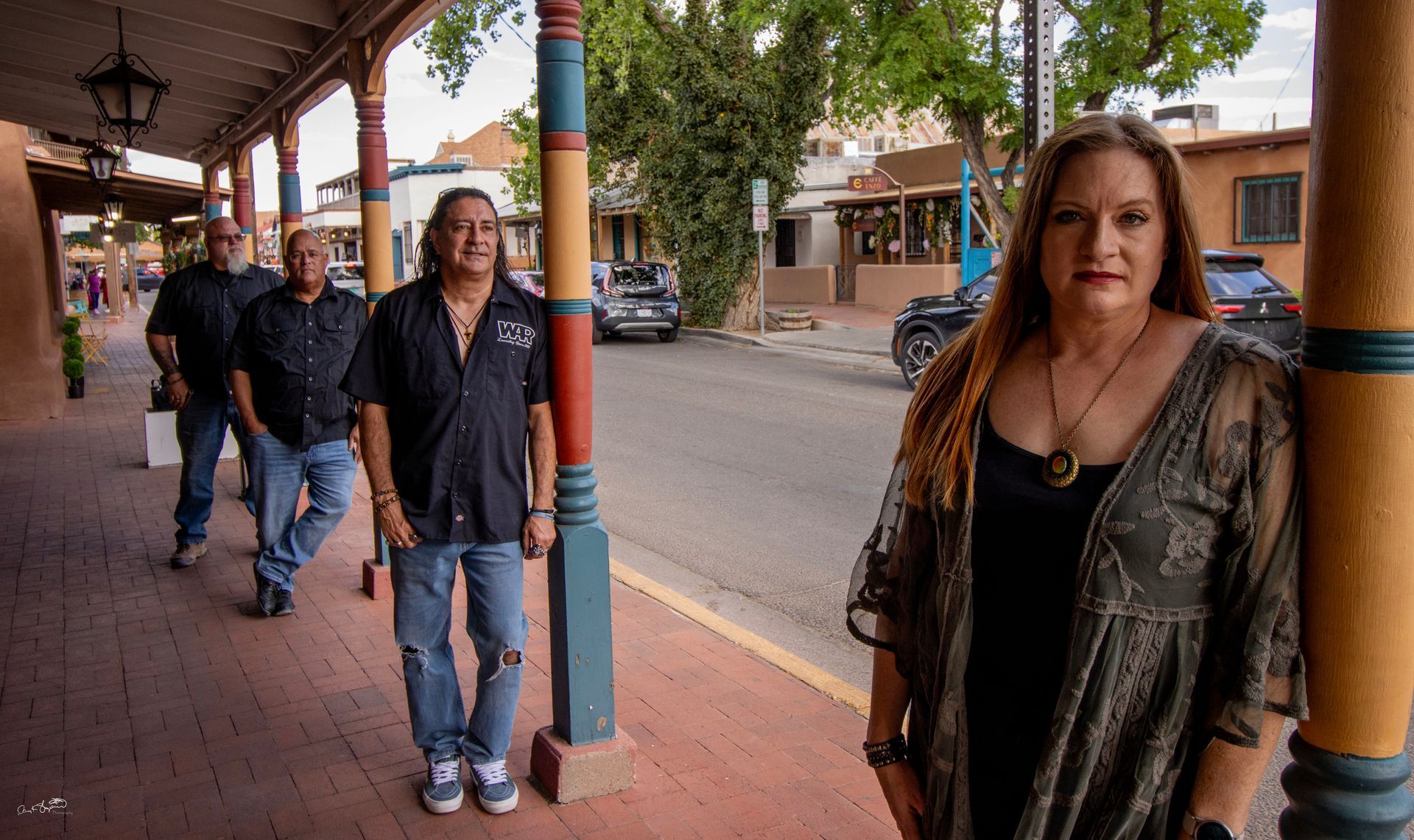A band of four stands on a sidewalk. A woman in a dark dress leans on a post. Street scene.