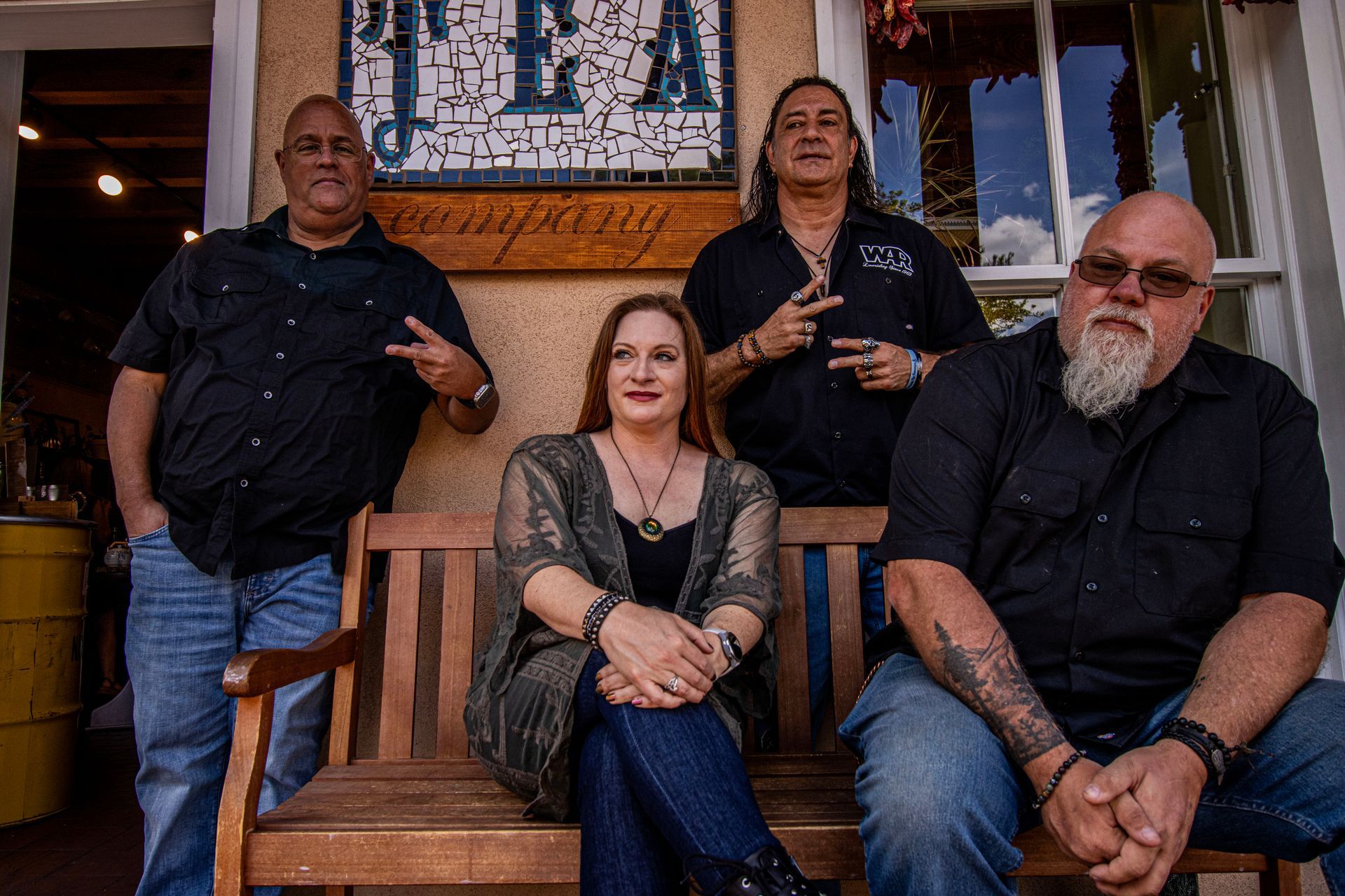 Four people posing outside, two men standing, two seated on a bench. All wearing black, some are making peace signs.
