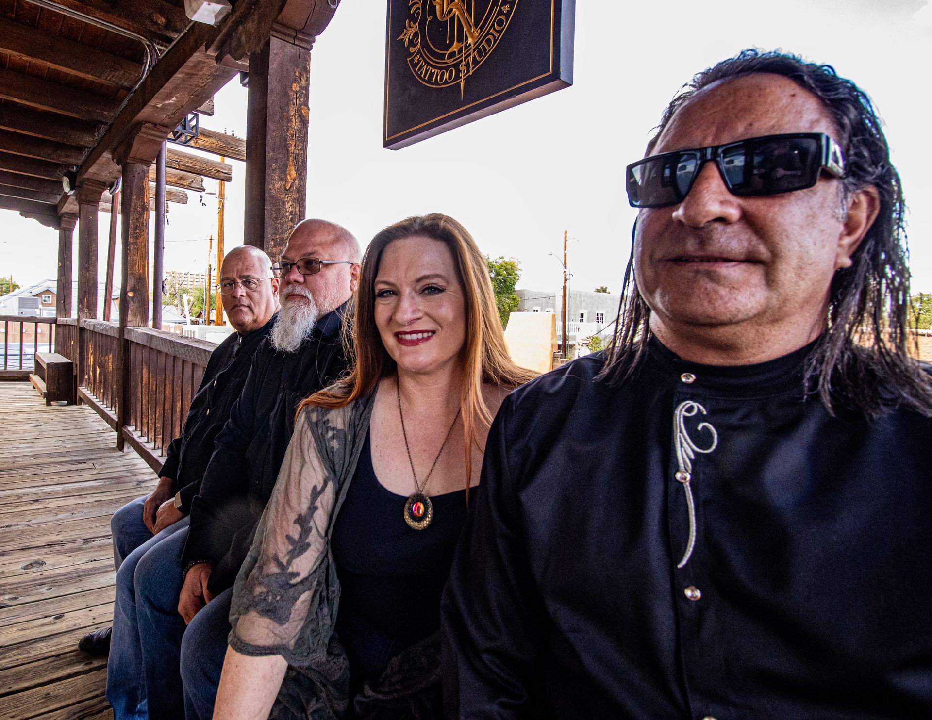 Band posing outdoors on a wooden porch; four people, a woman smiles, and one in sunglasses.
