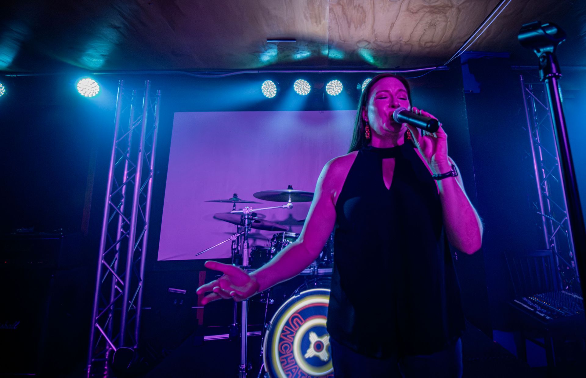 Woman singing into a microphone on stage, under blue and purple stage lights, drums in background.