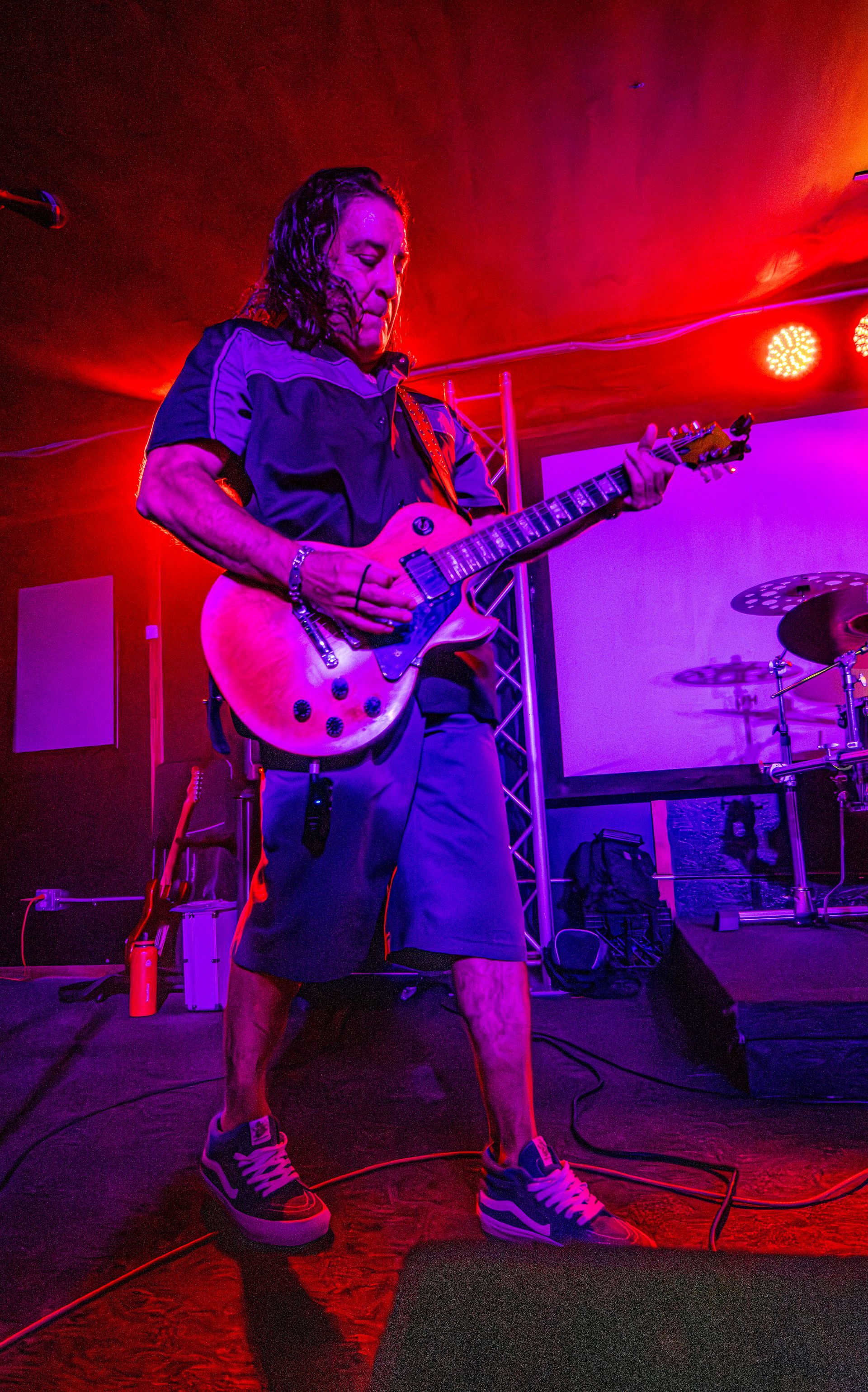 Guitarist on stage, lit with red and purple lights, playing a light-colored electric guitar.
