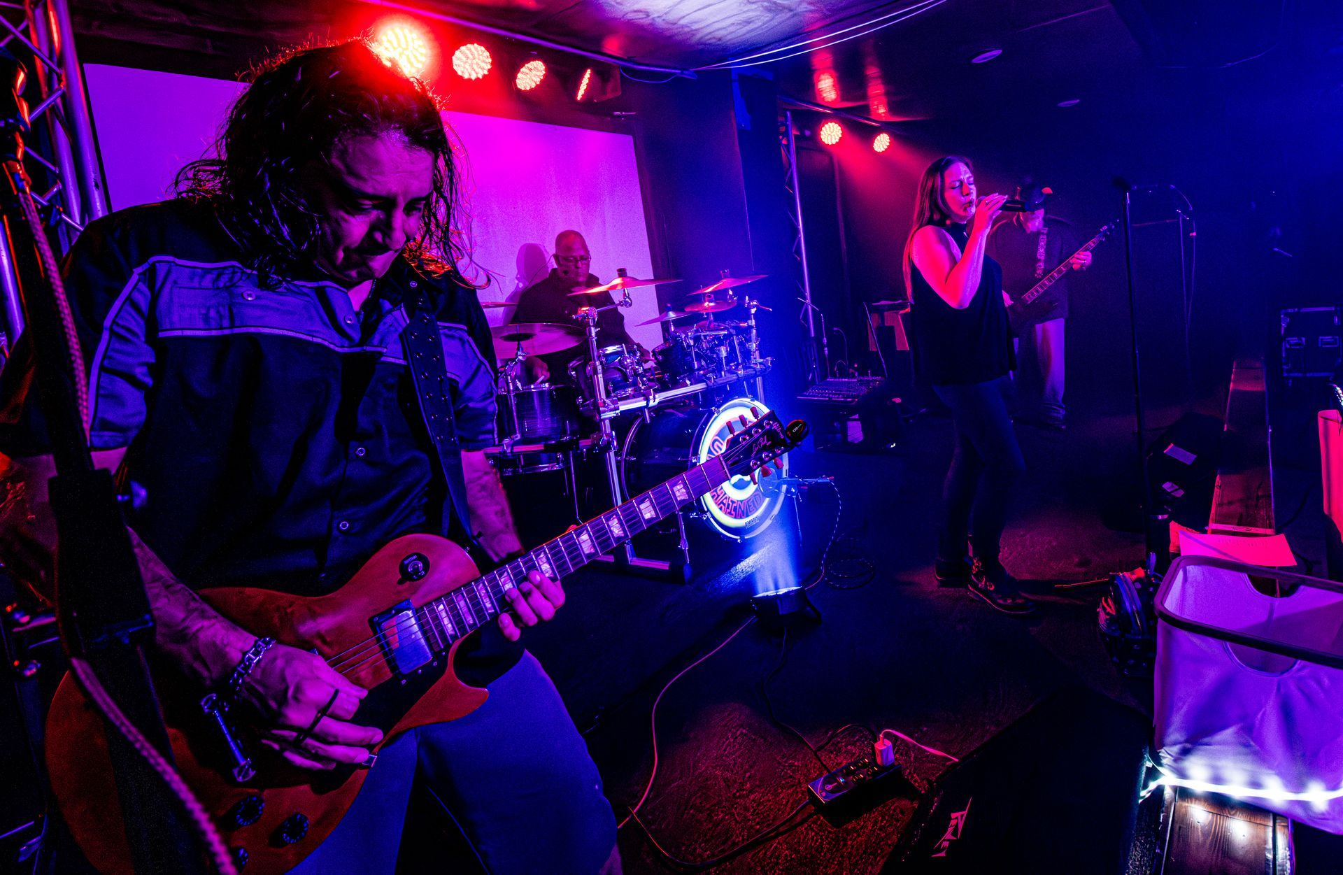 A band performing live on stage with a guitarist, singer, and drummer, under colorful stage lights.