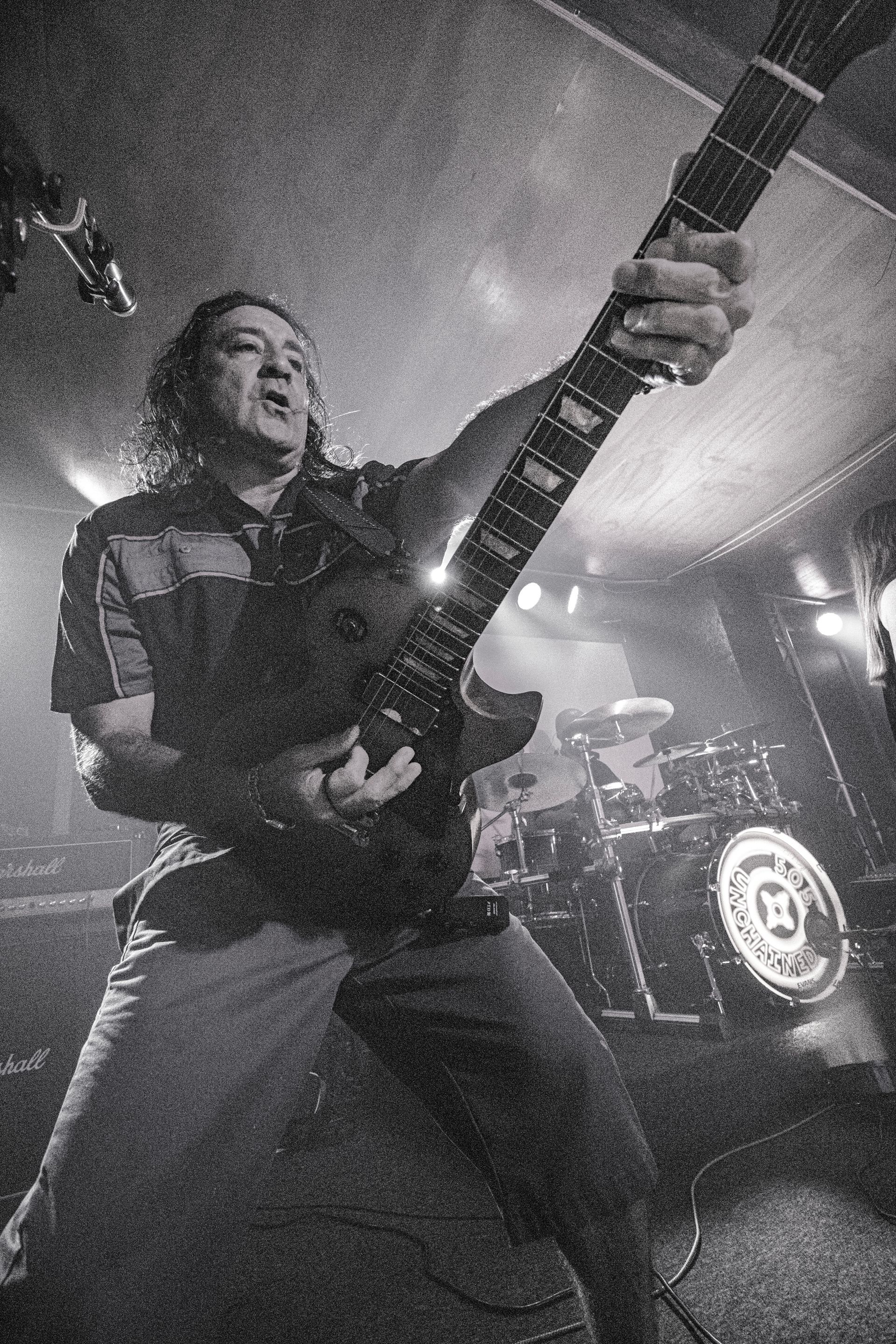 Man playing electric guitar onstage, raising it. Dark, grainy black and white photo.