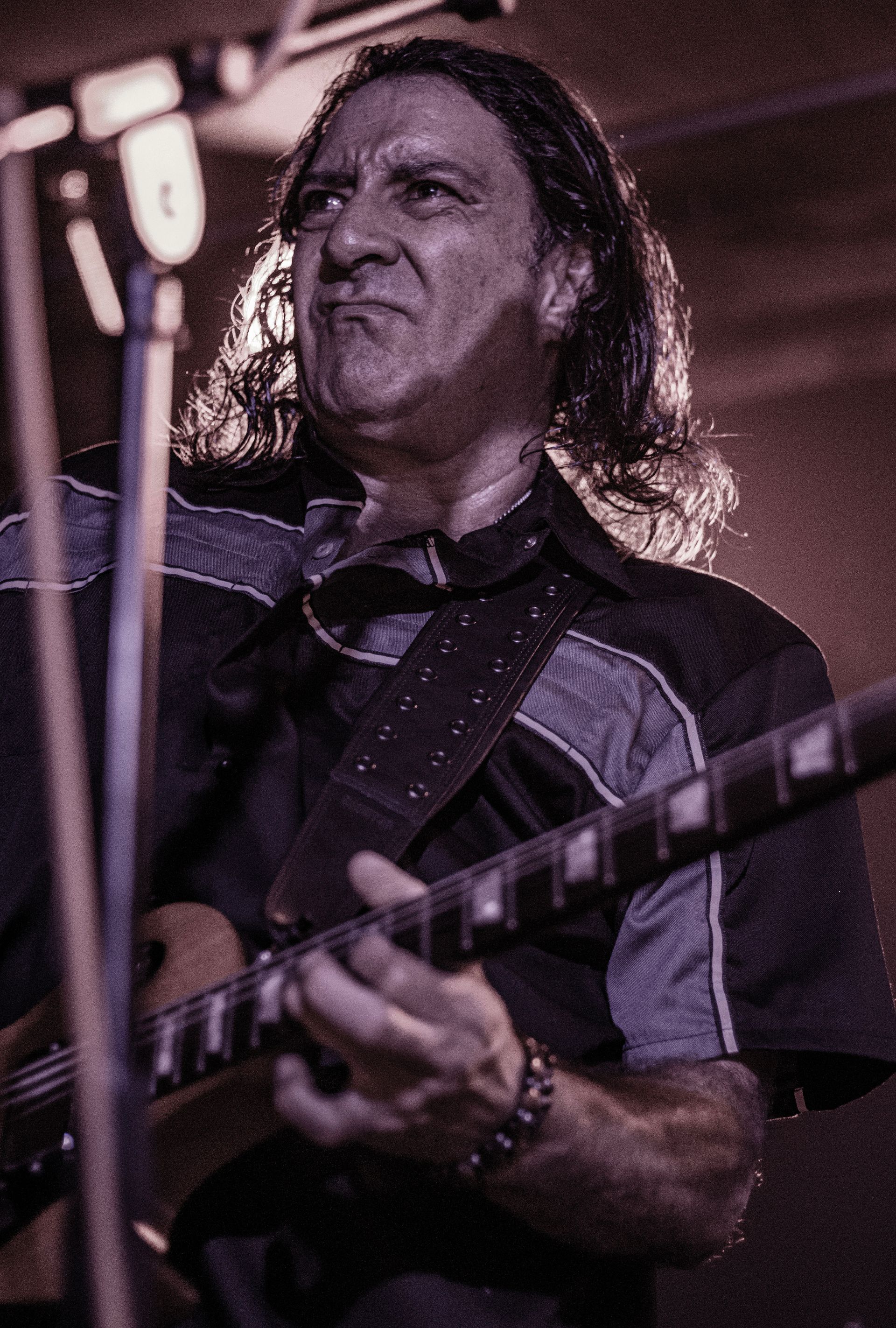 Man playing guitar onstage, furrowing brow, dark shirt, long hair, spotlight.