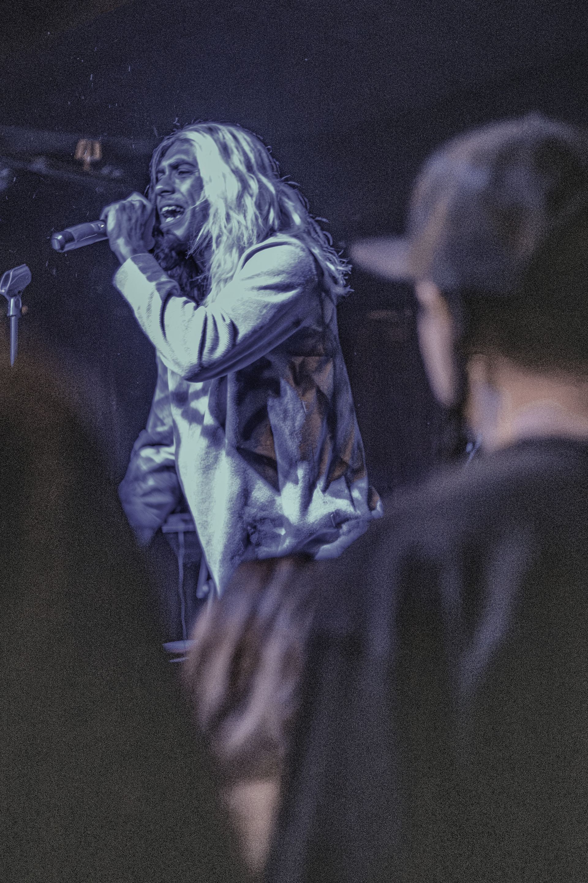 A person singing into a microphone onstage, lit in blue light, with long hair, seen from the waist up.