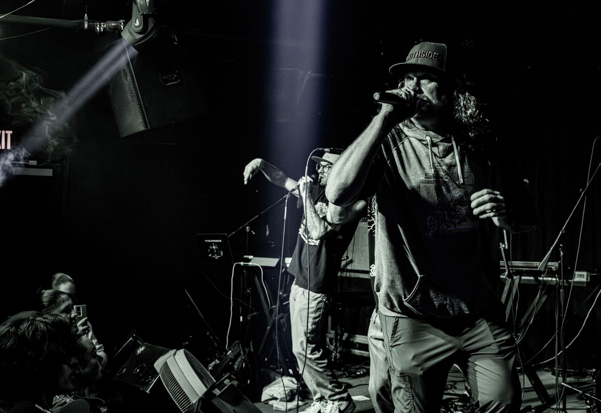 Musician on stage rapping into a microphone, wearing a hat, with another person in the background.