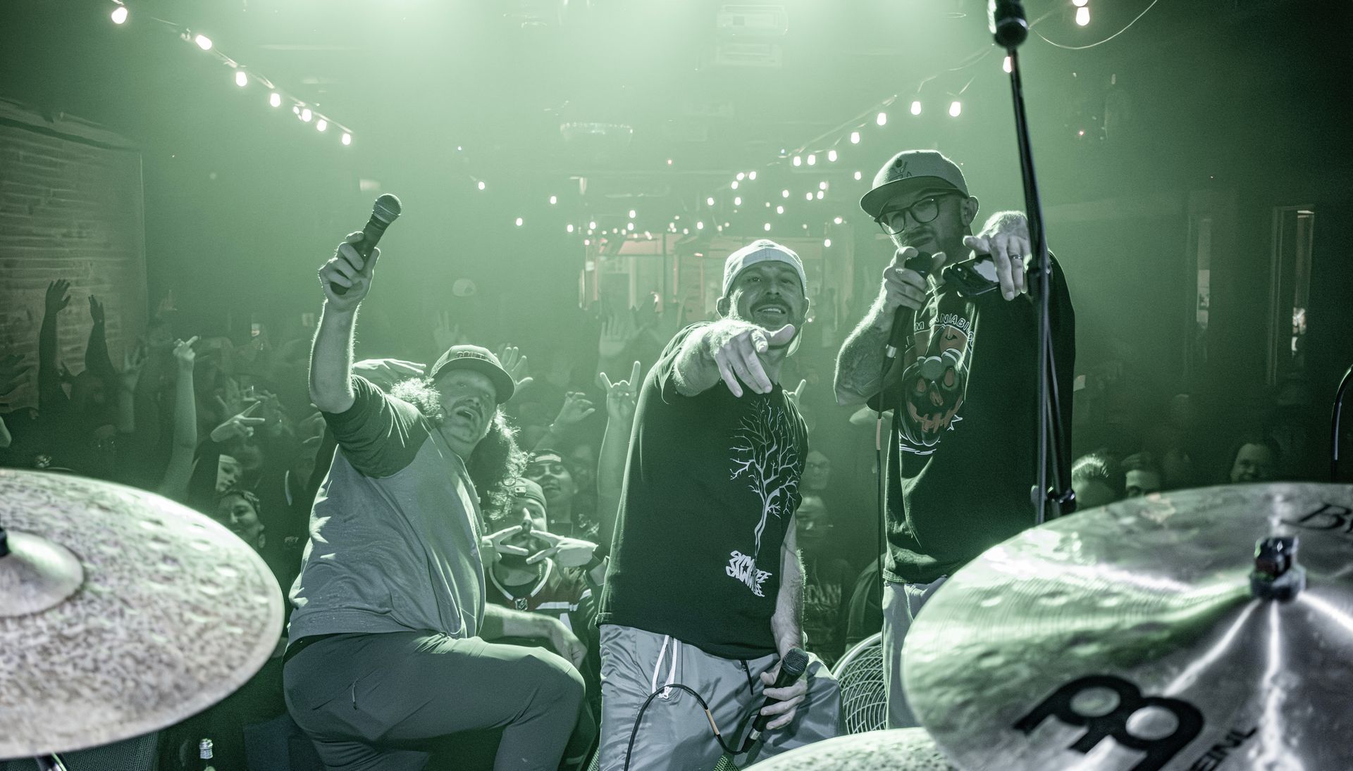 Band performing on stage, lit with green lights. Two singers point at the crowd. Drum set in the foreground.