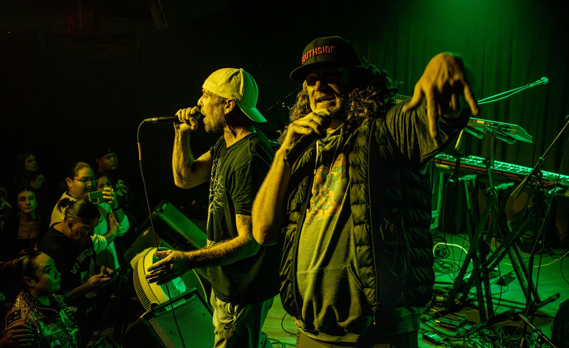 Two men on stage, rapping into microphones. Green lighting. Audience visible in background.