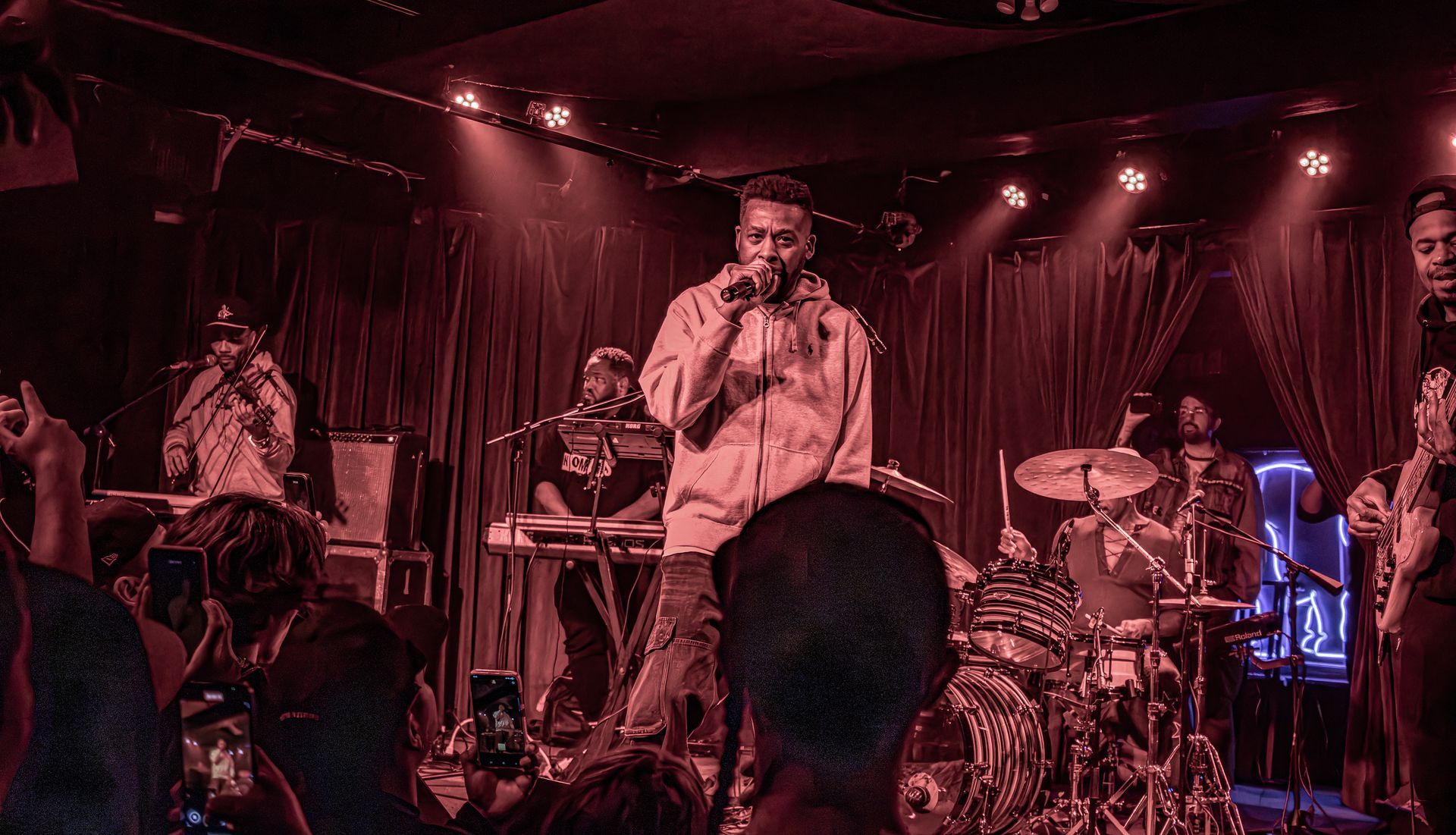 Band performing on stage under red lights; vocalist at microphone, crowd in front.