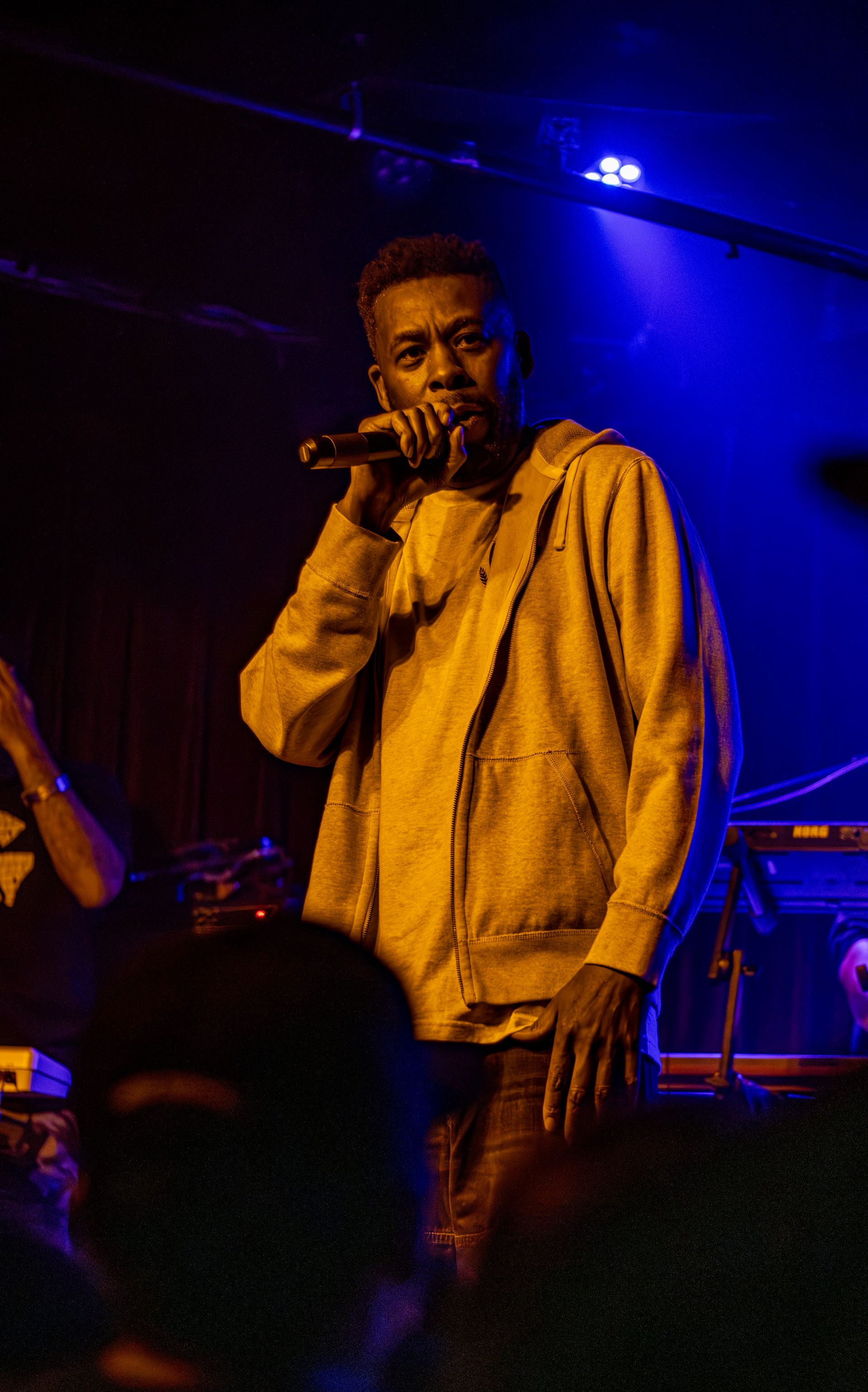 Man singing into a microphone on stage, wearing a yellow hooded sweatshirt, under blue stage lights.
