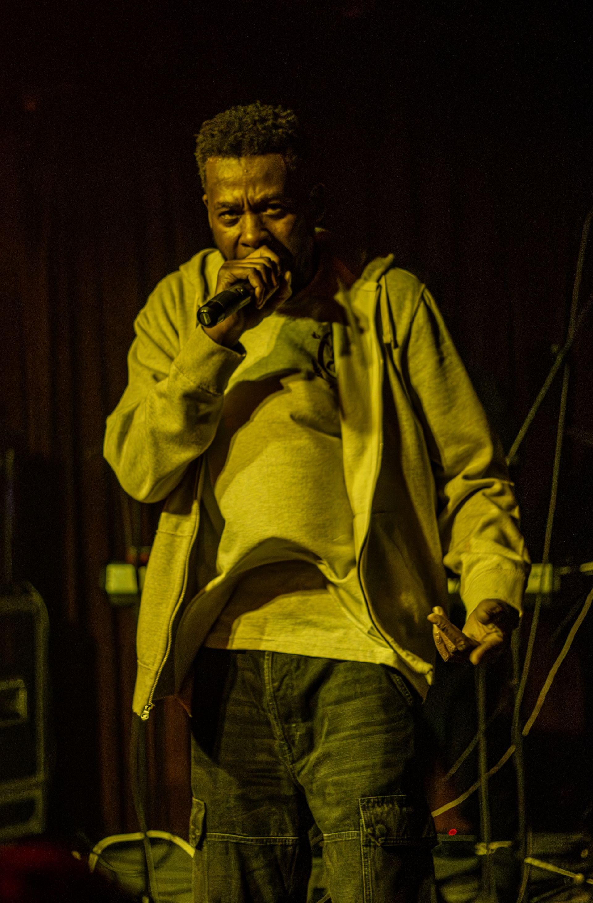 A person holding a microphone, performing on stage. They wear a light jacket and jeans, lit with yellow stage light.