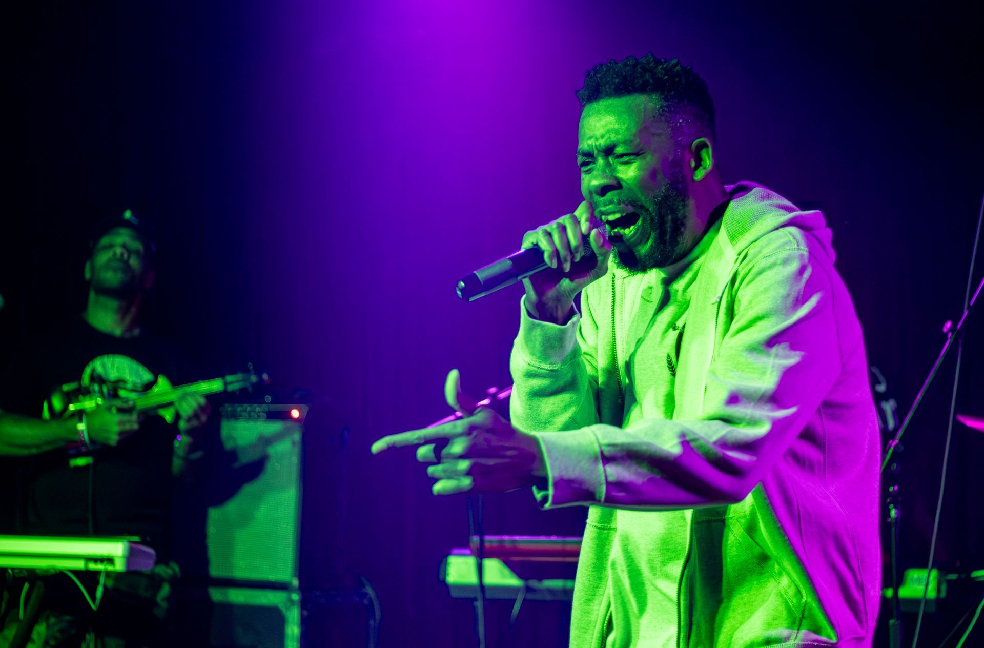 Man with microphone, performing on stage with green and purple lighting.