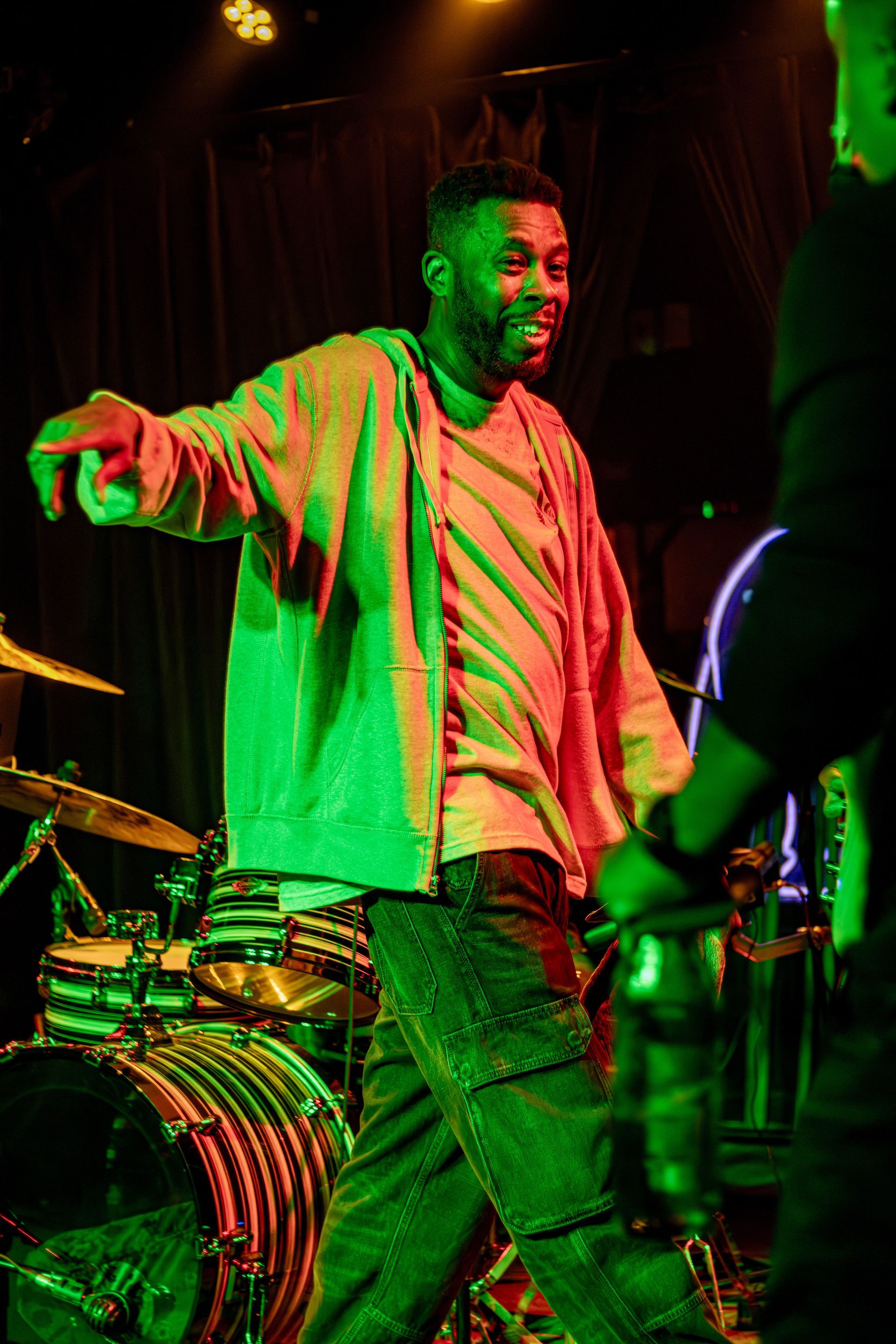 Man in light-colored hoodie dancing on stage with drums in the background, lit with green and red light.