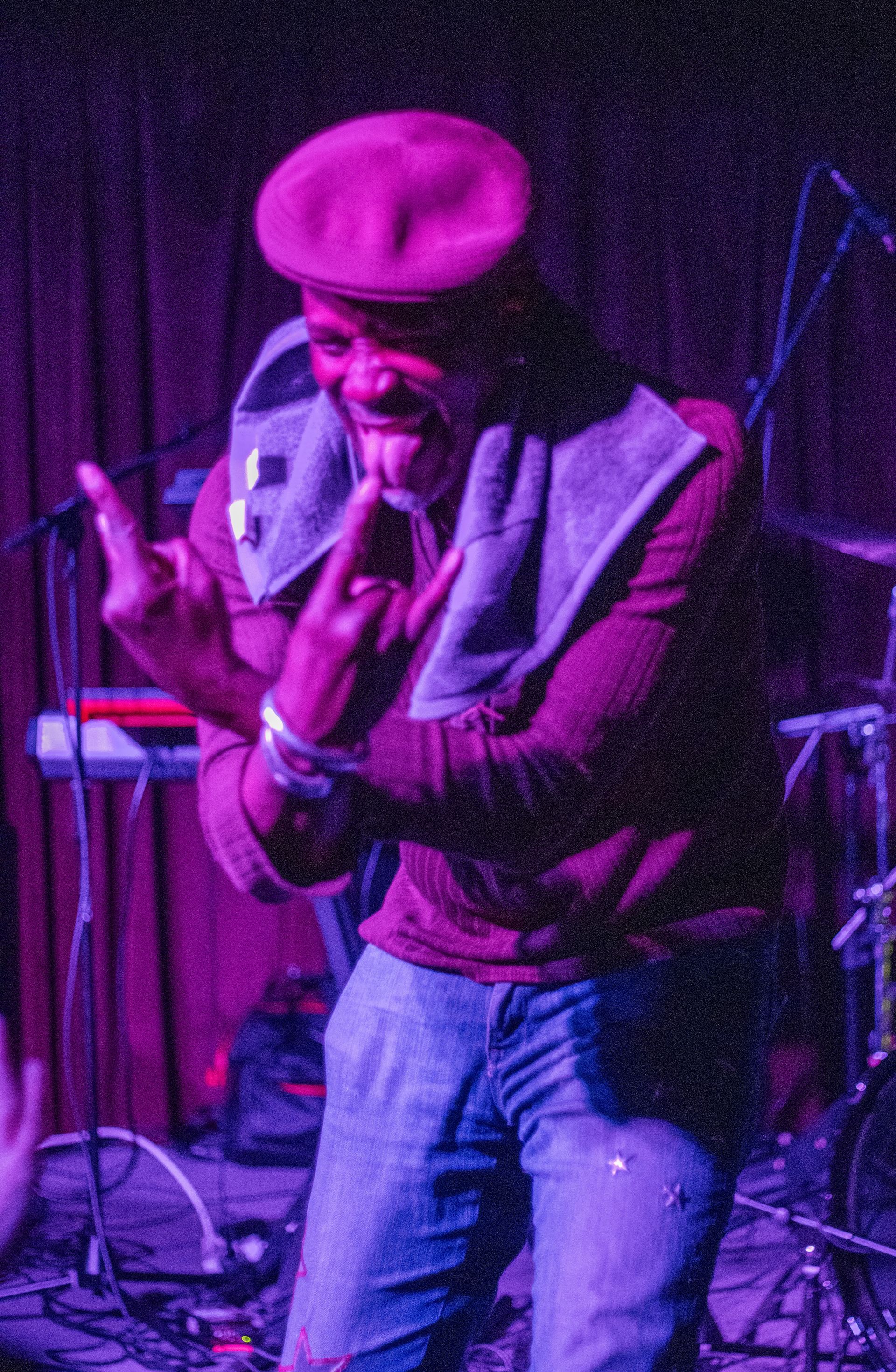 Person on stage wearing a maroon beret and sweater, denim pants, gesturing with hands. Purple lighting.