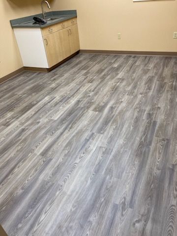 Gray wood-look flooring in a room with a sink and cabinet. Beige walls and a brown baseboard are visible.