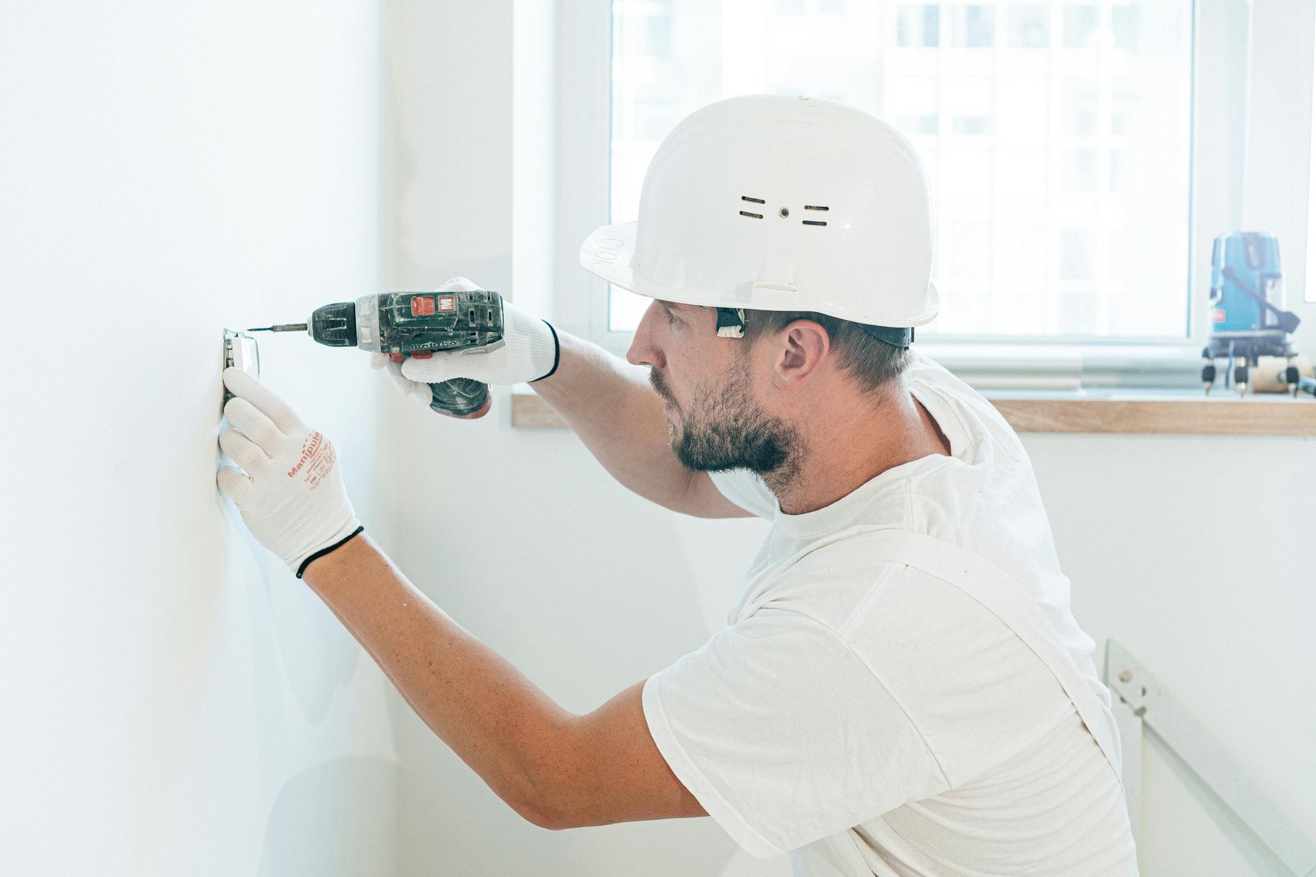 An electrician in white drilling a whole, with a power drill, into a while wall in a brightly lit room