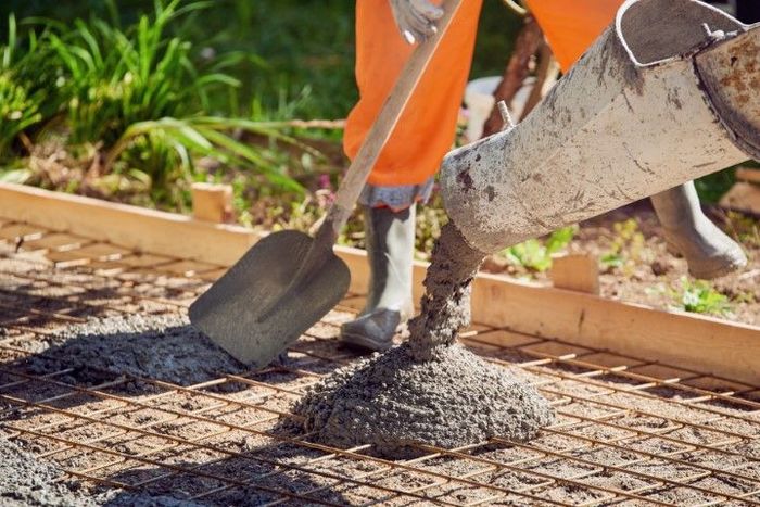 Concrete being poured from a chute onto a wire mesh, with a person holding a shovel, in an outdoor setting.