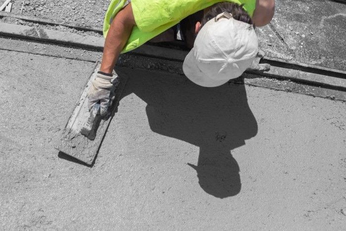 Person in a neon yellow shirt and white hat smoothing wet concrete with a trowel.