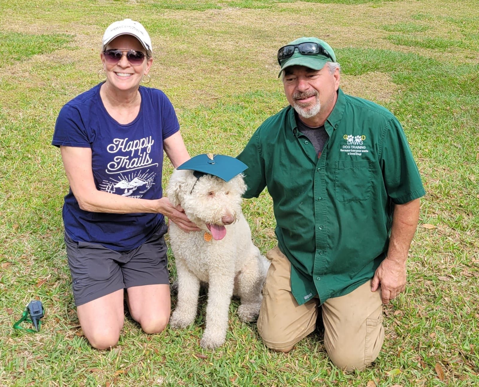 A man and a woman are kneeling next to a dog wearing a graduation cap.