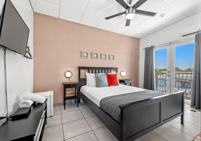 Bedroom with bed, TV, and balcony door. Features a black bed frame, pink walls, and bright natural light.