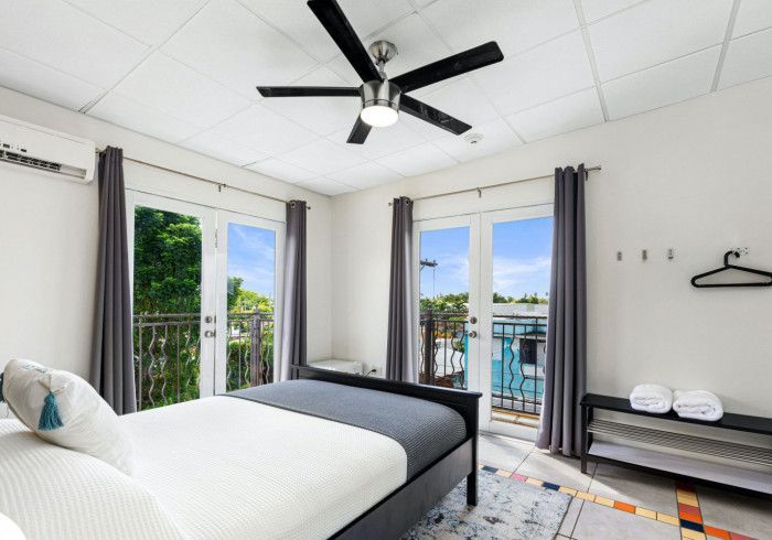 Bedroom with bed, balcony doors, ceiling fan, and gray curtains; a blue sky visible.
