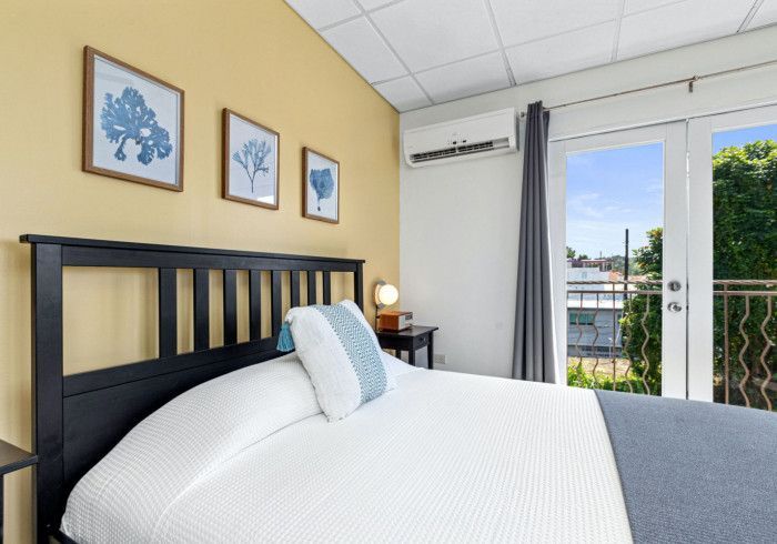 Bedroom with a black bed frame, white bedding, and a balcony door. Yellow wall with blue artwork.