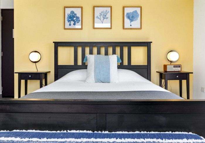 Bedroom with a yellow wall, black bed frame, and three blue-framed artworks above.